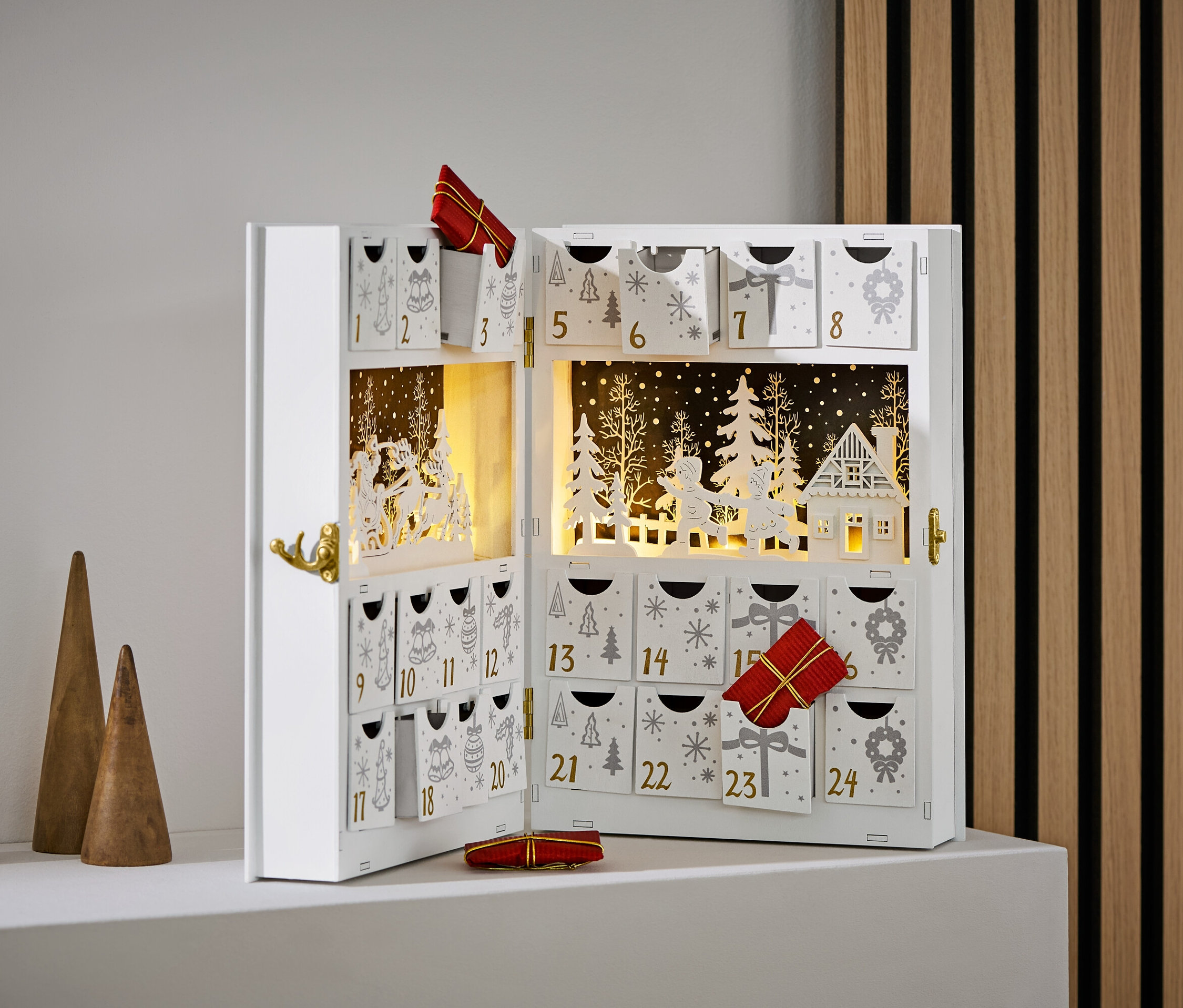 Otwarty, biały kalendarz adwentowy z ozdobami i prezentami, obok trzech drewnianych stożków.