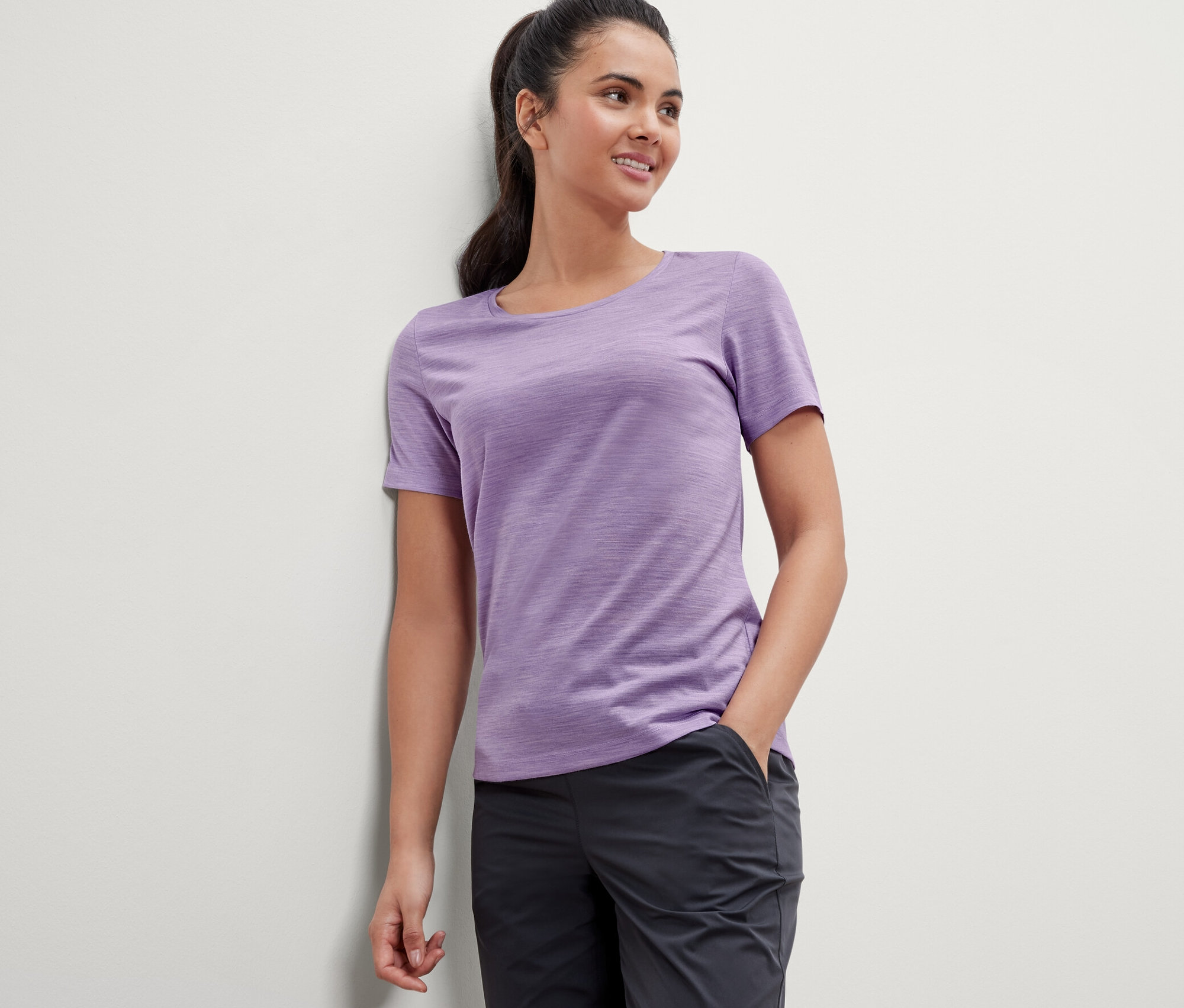 Kobieta oparta o ścianę ma na sobie liliową koszulkę z wełny merino.
