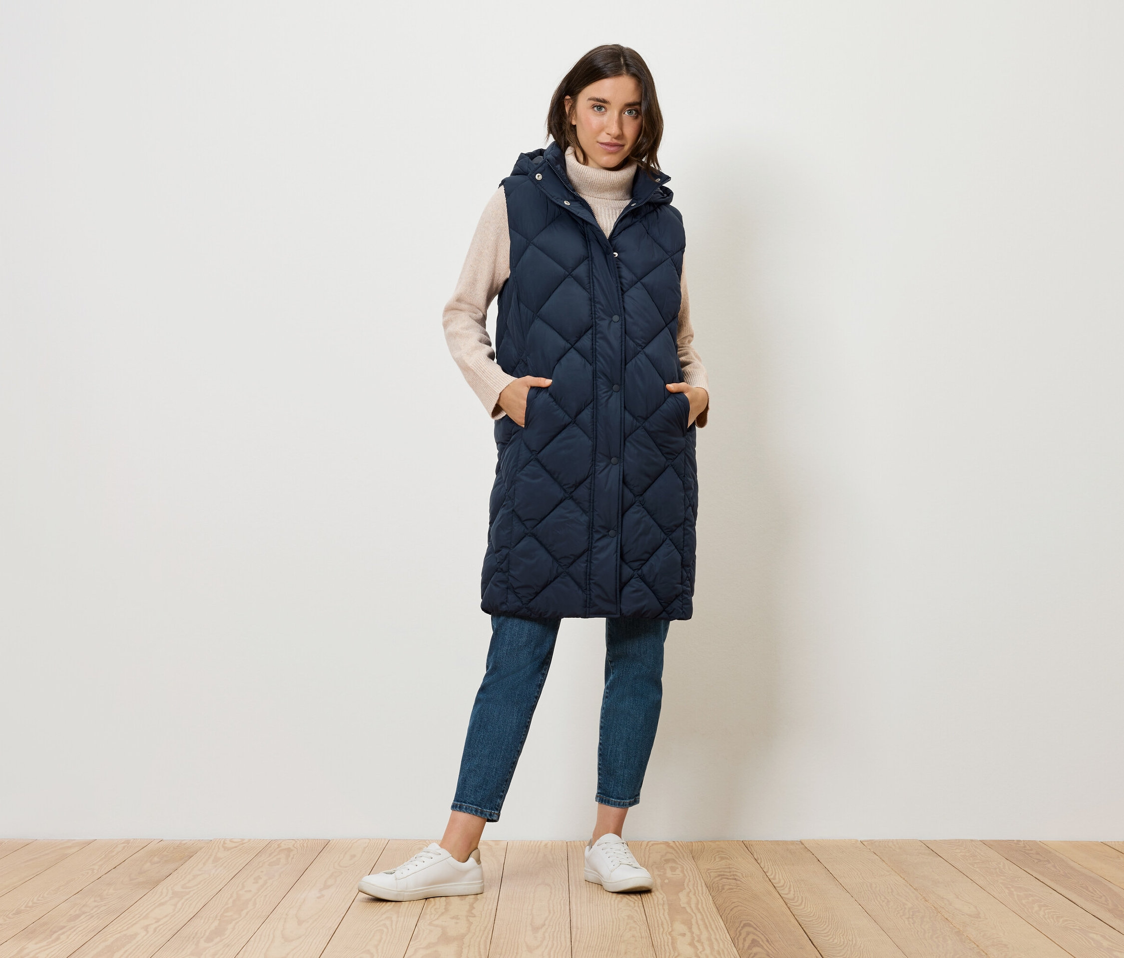 Kobieta stoi i ma na sobie długą, pikowaną, granatową kamizelkę z kapturem, dżinsy i tenisówki.