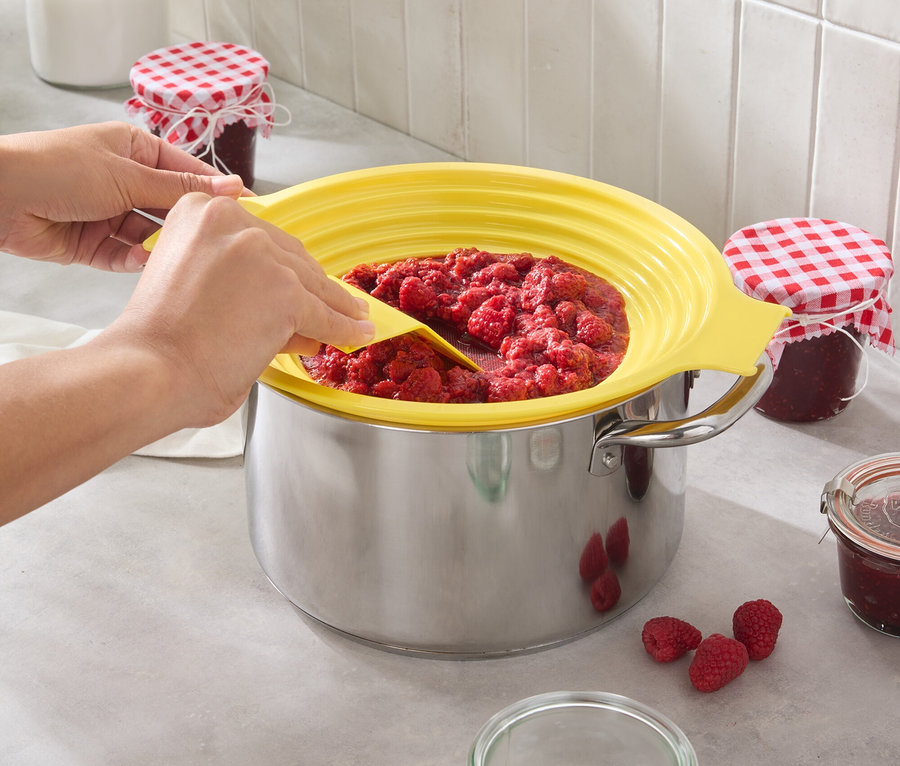 Osoba miesza dżem malinowy w garnku z żółtą osłoną przed pryskaniem. Kilka malin i słoików stoi na blacie kuchennym.