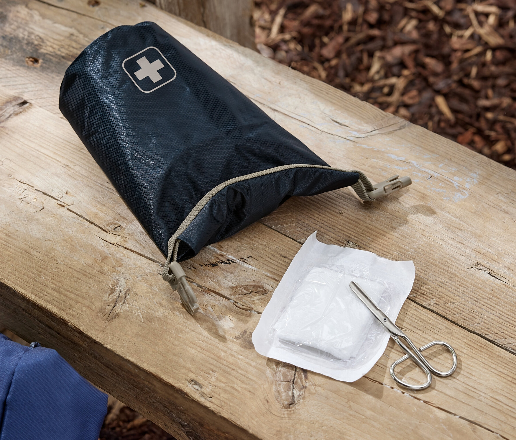 Na drewnianej ławce leży otwarta apteczka pierwszej pomocy, bandaż i nożyczki.