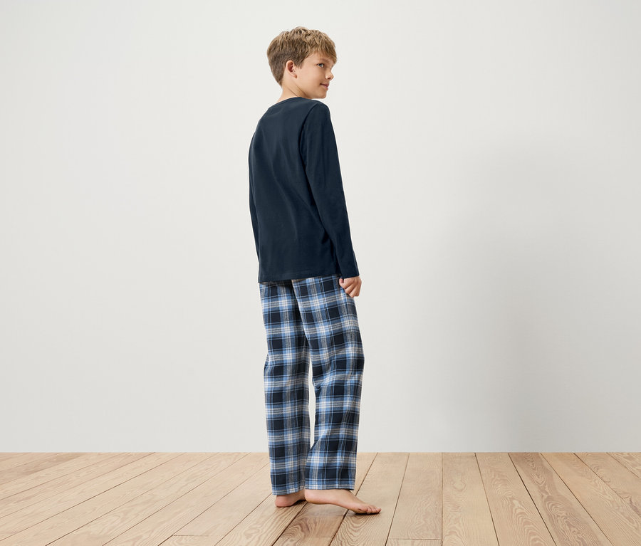Chłopiec w granatowej piżamie z długim rękawem i niebieskich spodniach od piżamy w kratę stoi boso na drewnianej podłodze.