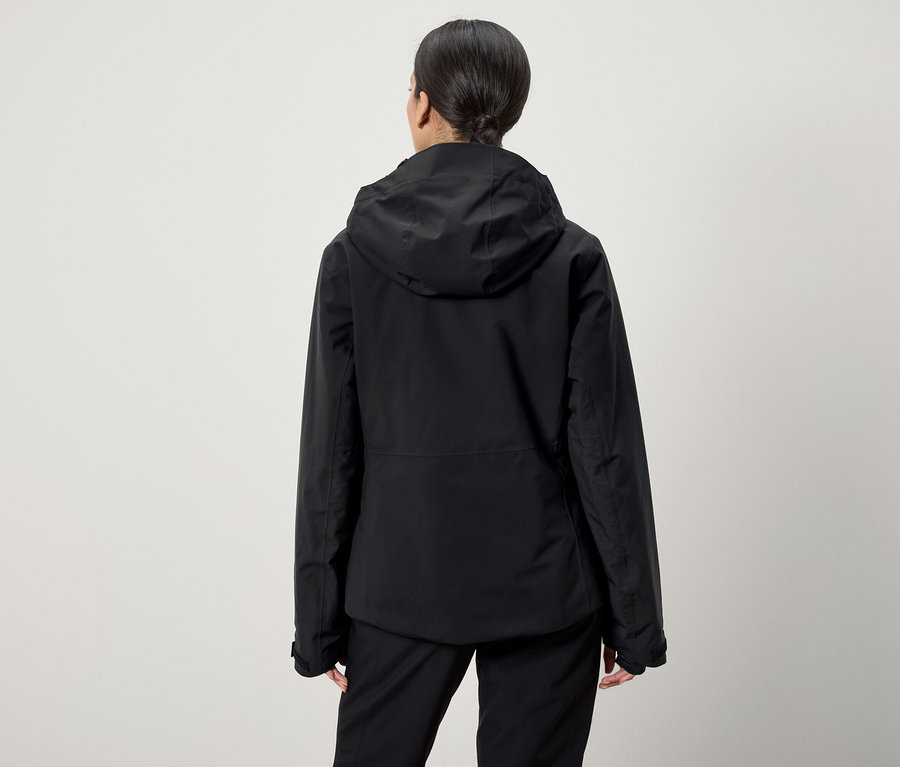 Z tyłu widoczna jest kobieta z włosami związanymi w koński ogon, ubrana w czarną kurtkę z kapturem i czarne spodnie.