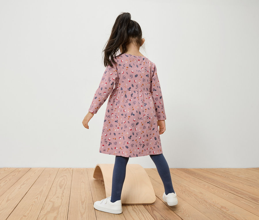 Dziewczynka w kwiecistej sukience stoi na drewnianej desce do balansowania.