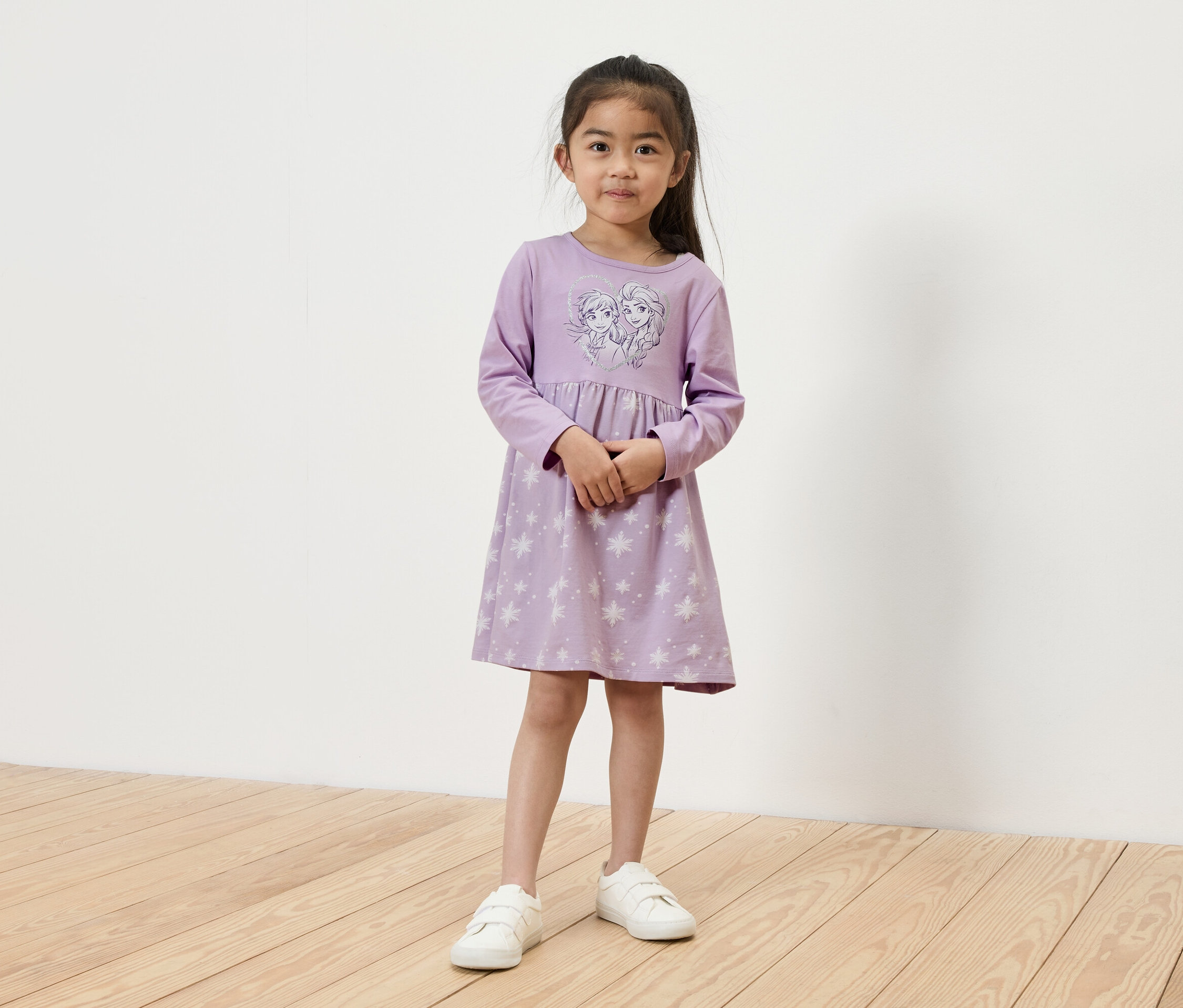 Dziewczynka stoi i ma na sobie jasno fioletową sukienkę z długim rękawem z motywem Elsy i Anny oraz białe tenisówki.
