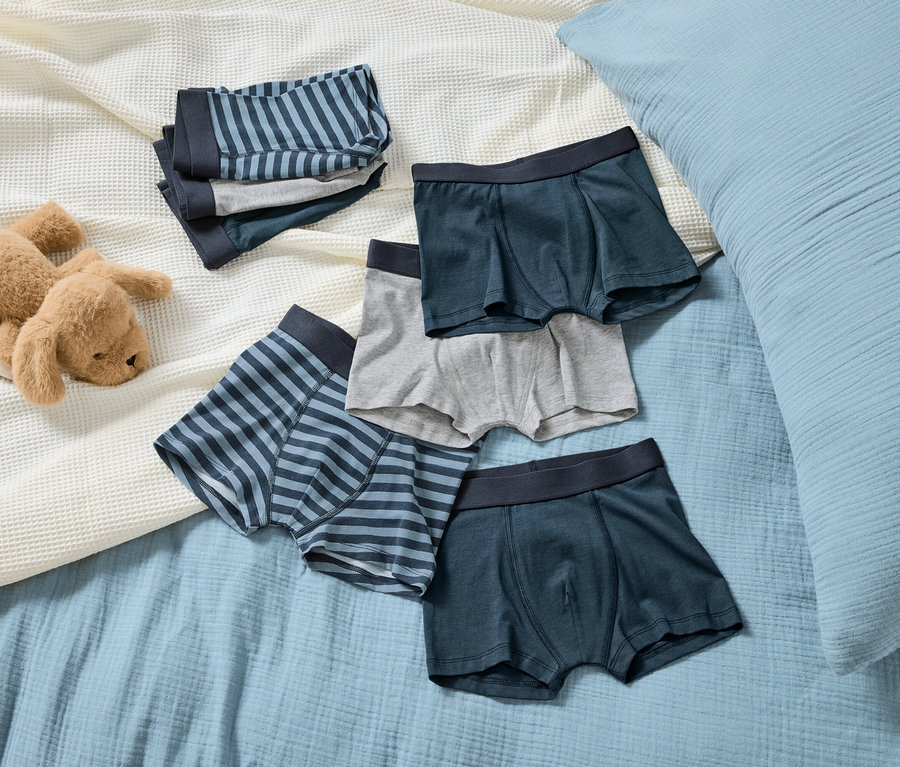 Na jasnoniebieskim kocu leży brązowy pluszowy pies obok stosu chłopięcych bokserek w paski i jednolitych kolorach.