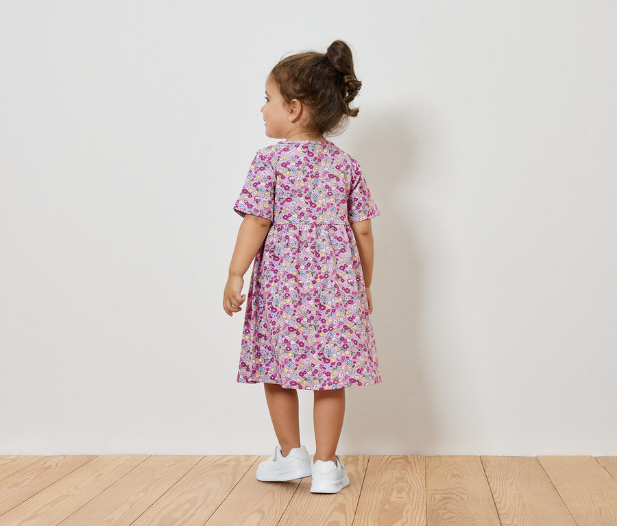 Dziewczynka w kwiecistej sukience i białych tenisówkach stoi tyłem do kamery.