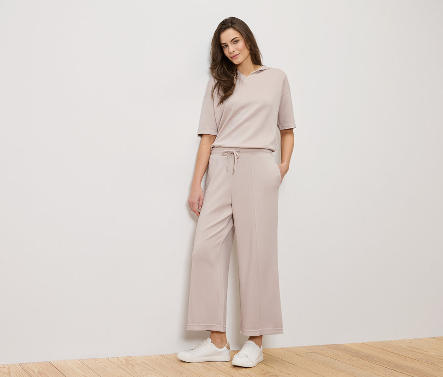 Kobieta stoi na tle białej ściany, ubrana w beżowy strój i białe tenisówki.