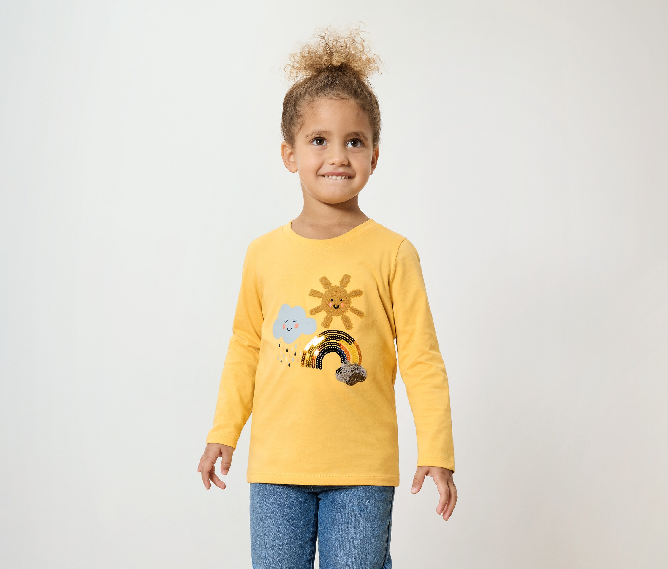 Dziewczynka z kręconymi włosami ma na sobie żółtą koszulkę z długim rękawem z chmurą, słońcem i tęczą oraz niebieskie dżinsy.