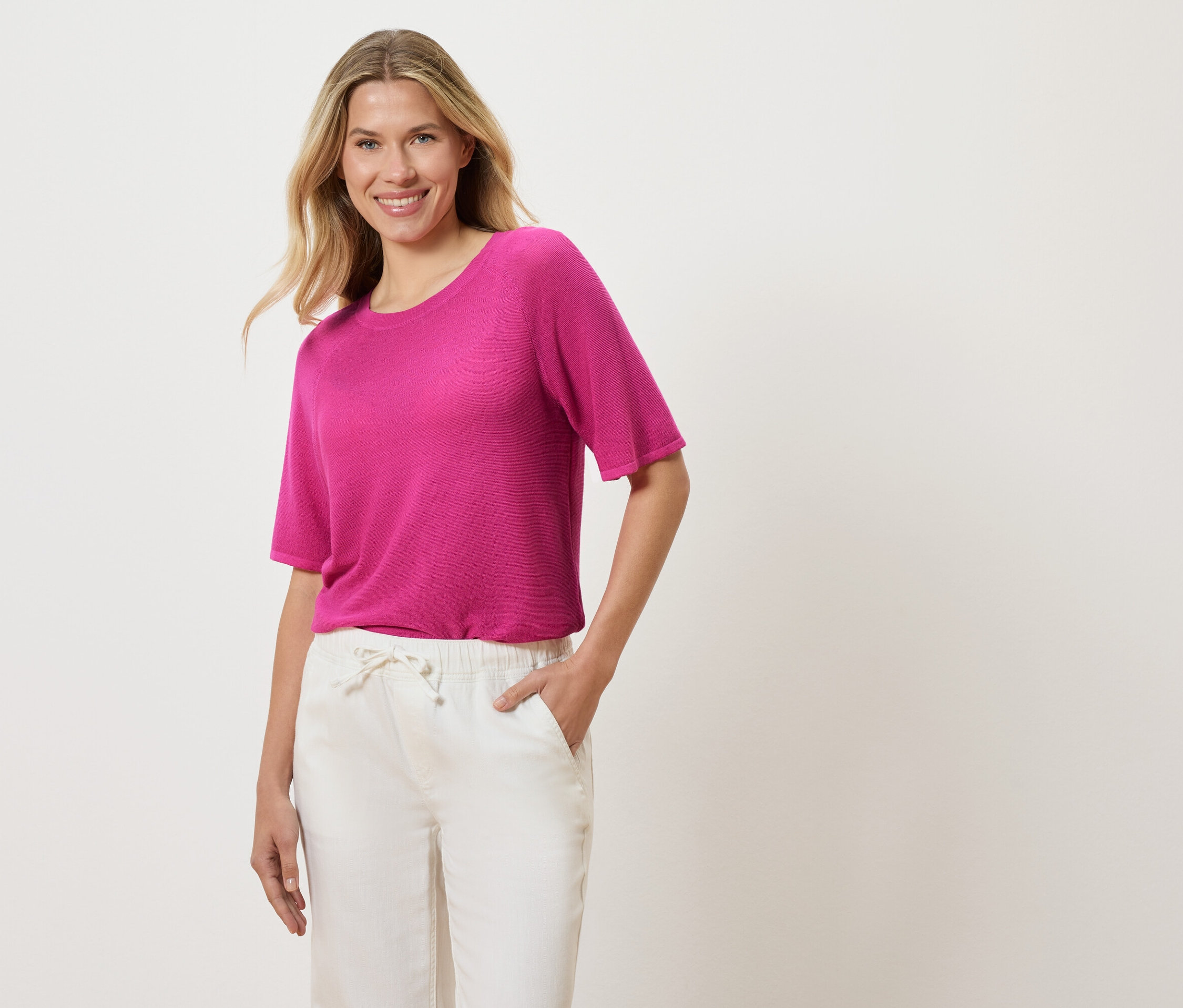 Kobieta o blond, falistych włosach ma na sobie różowy T-shirt i białe spodnie, trzyma rękę w kieszeni.