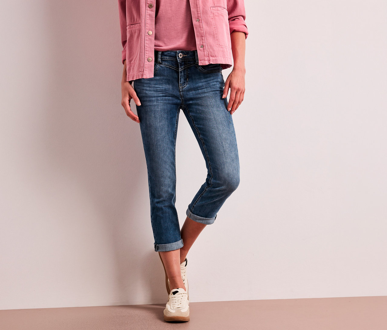 Osoba w różowej kurtce, różowym topie, niebieskich podwiniętych jeansach 7/8 Street One „Jane” i jasnych sneakersach.