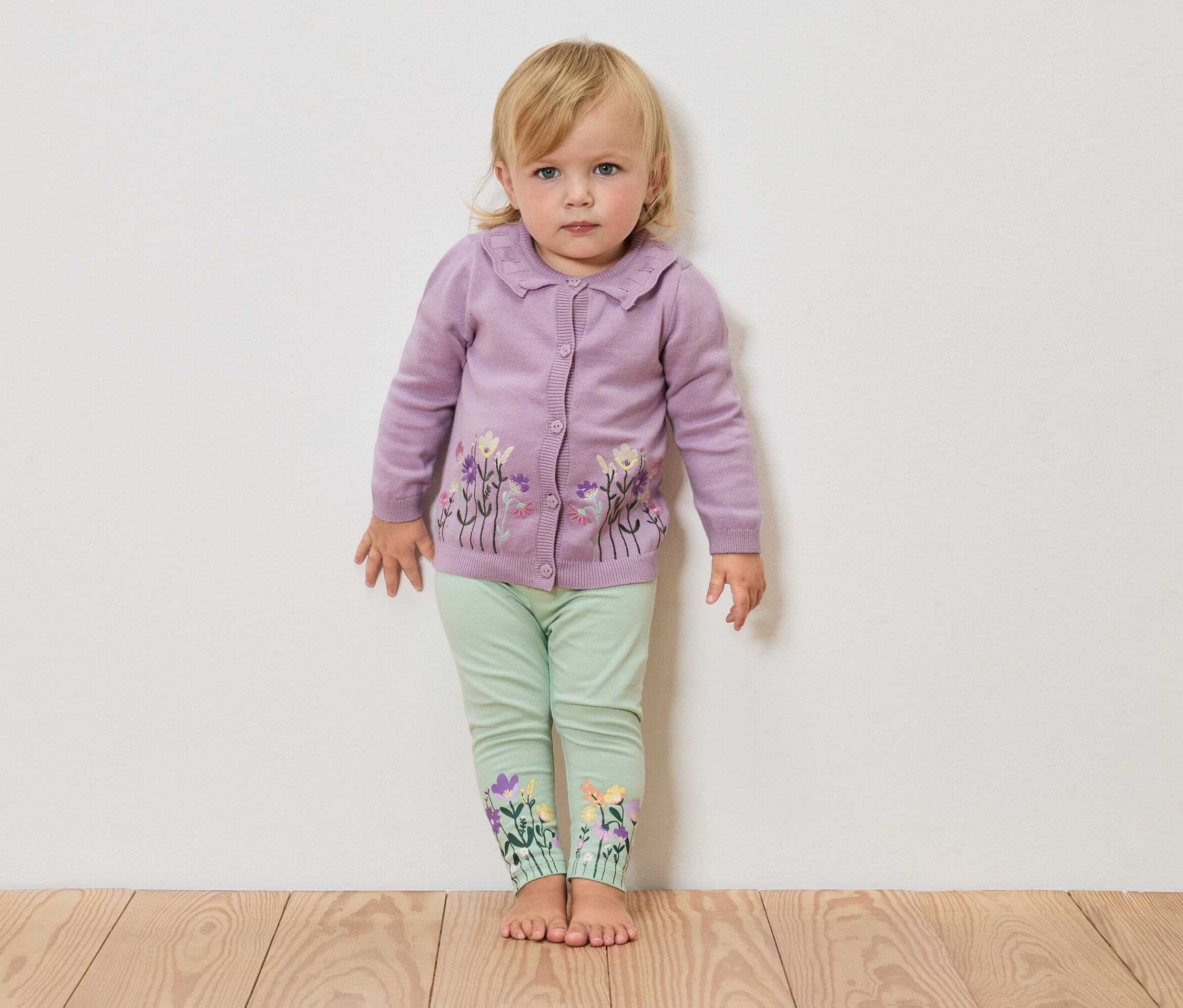 Blondwłosa dziewczynka stoi boso na drewnianej podłodze na tle białej ściany, ma na sobie liliowy kardigan w kwiaty i zielone legginsy z kwiatami na nogawkach.
