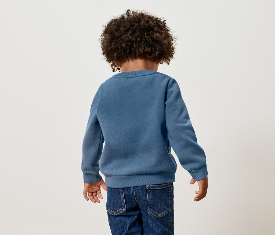 Widok z tyłu dziecka z kręconymi włosami, ubranego w niebieski sweter i dżinsy.
