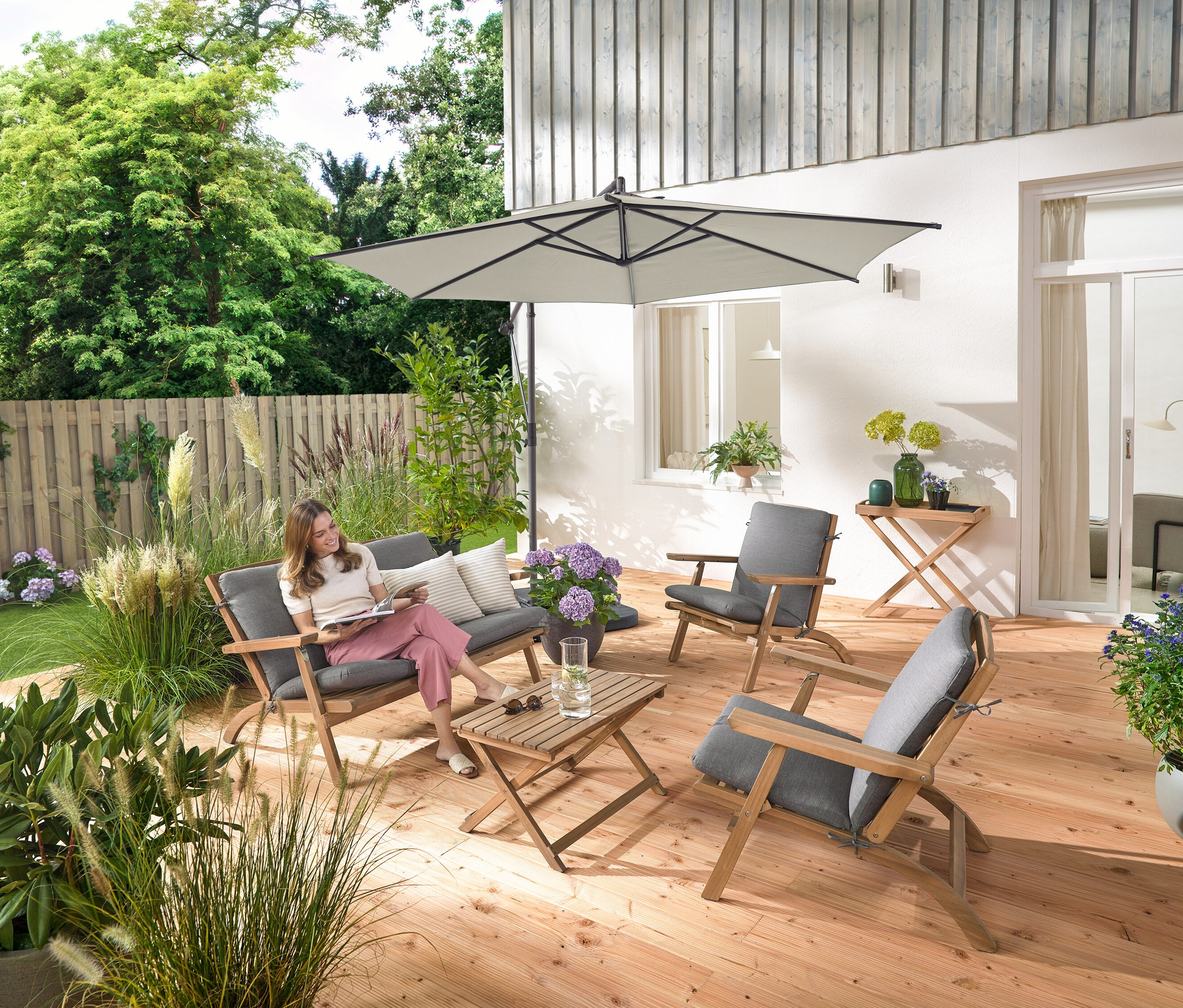 Kobieta siedzi na składanym zestawie wypoczynkowym ogrodowym z poduszkami Premium, czyta książkę i jest otoczona przez stół ogrodowy i 2 poduszki zewnętrzne z lnu.