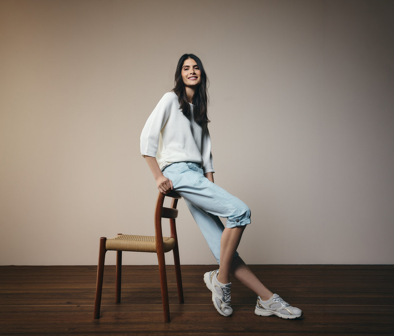 Uśmiechnięta kobieta opiera się o krzesło, ma na sobie jasnoniebieskie spodnie 3/4 „New York” CECIL i biały sweter.