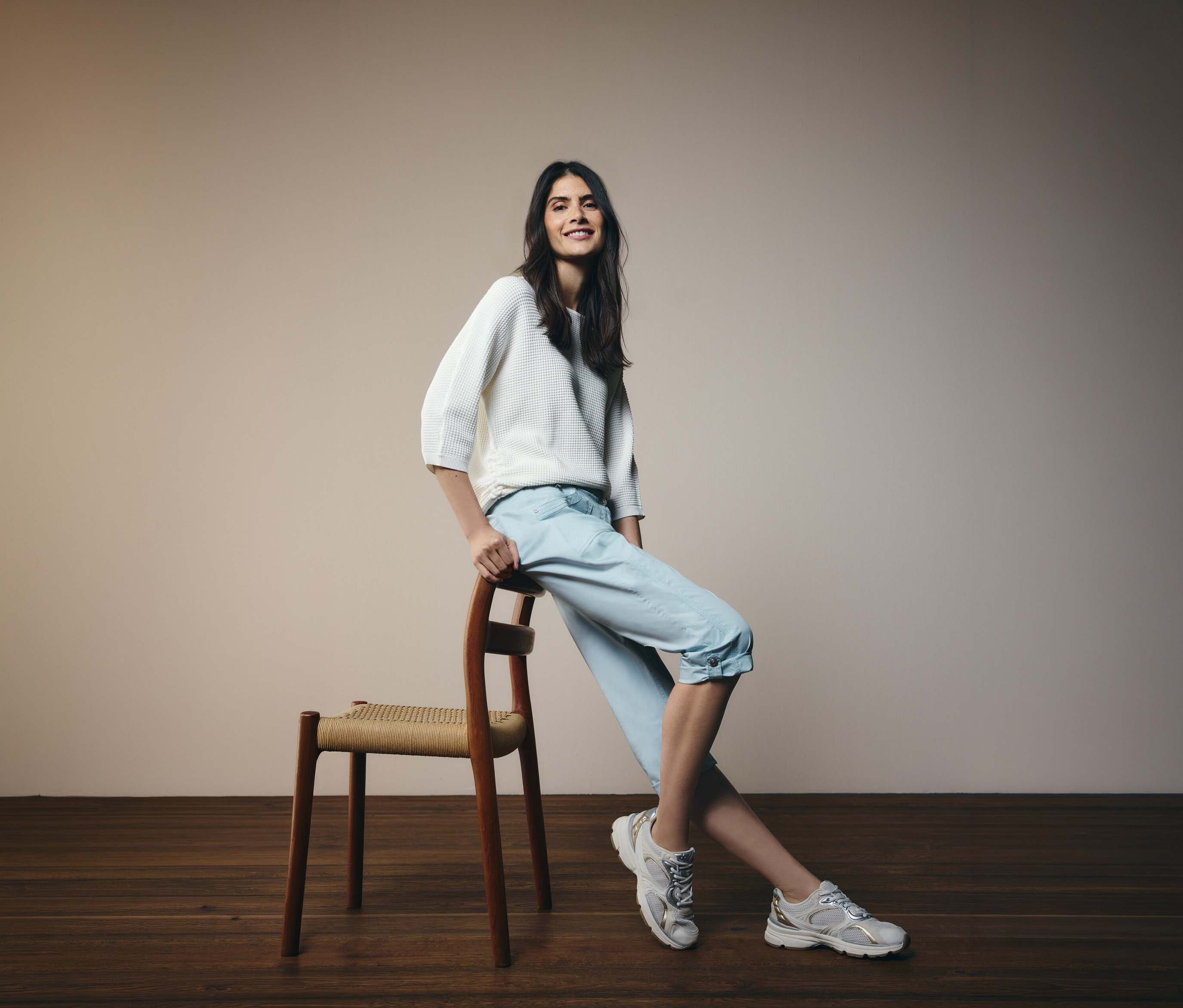 Uśmiechnięta kobieta opiera się o krzesło, ma na sobie jasnoniebieskie spodnie 3/4 „New York” CECIL i biały sweter.