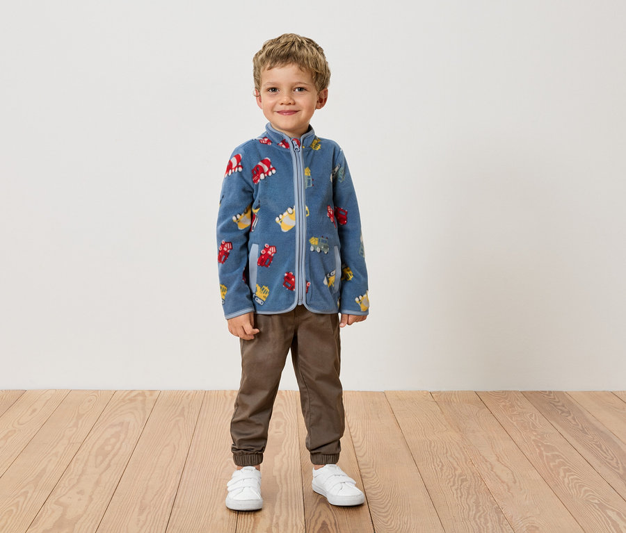 Chłopiec stoi na tle ściany w niebieskiej bluzie polarowej we wzory pojazdów, brązowych spodniach i białych butach.