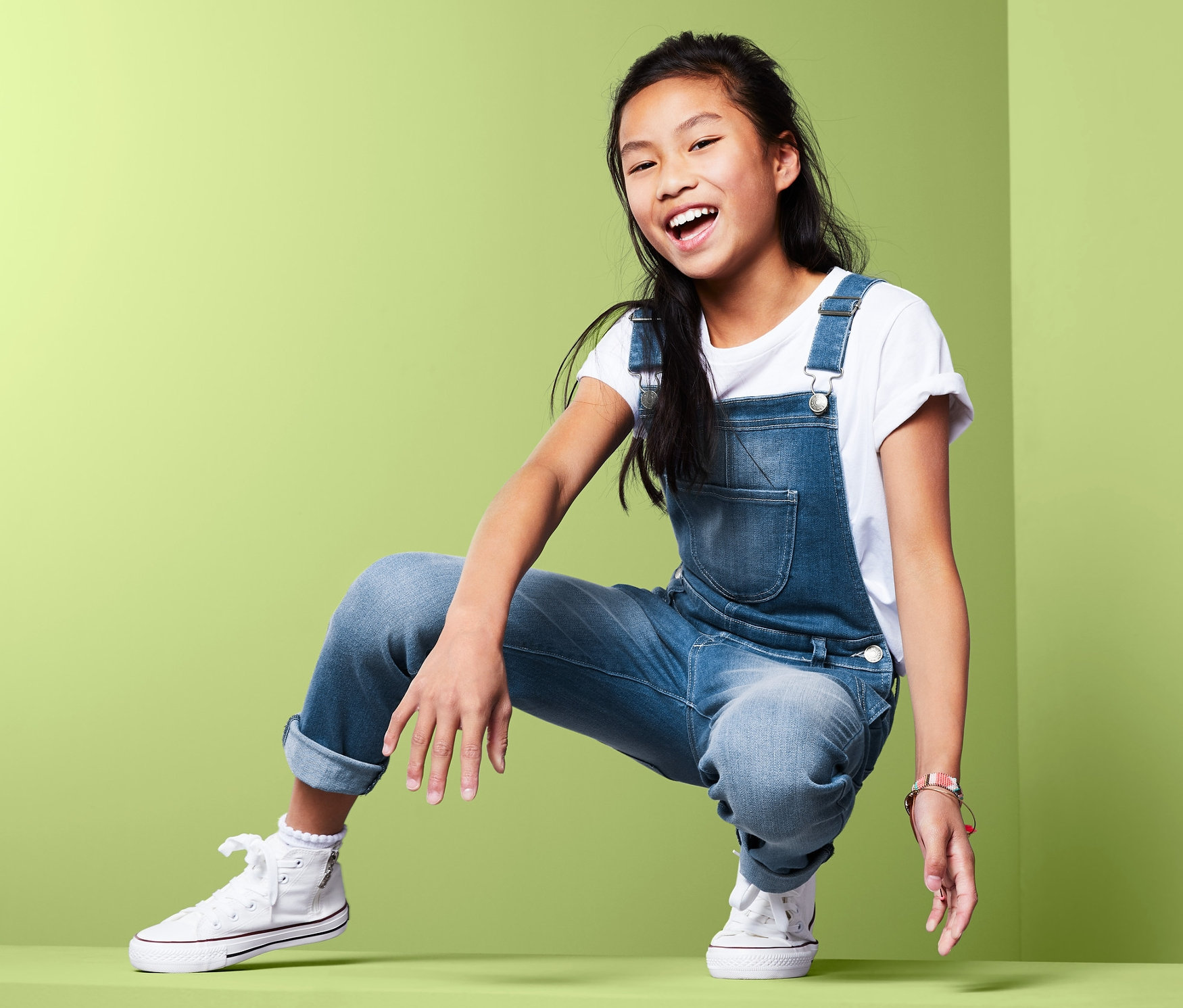Uśmiechnięta Azjatka w dżinsowych ogrodniczkach dziewczęcych z długimi nogawkami pozuje w kucki na zielonym tle.
