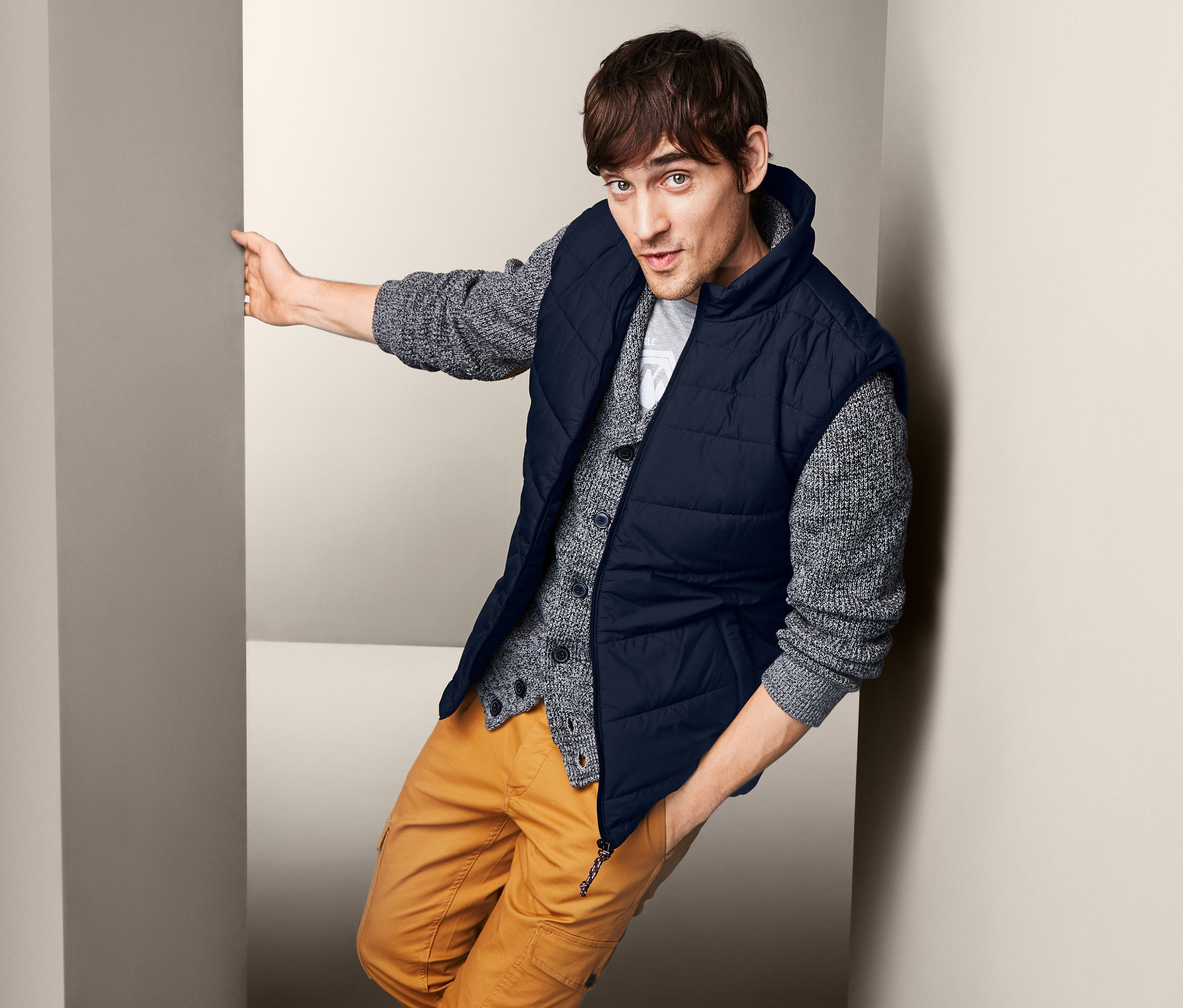 Mężczyzna opiera się o ścianę i ma na sobie męską kamizelkę pikowaną, sweter i spodnie.