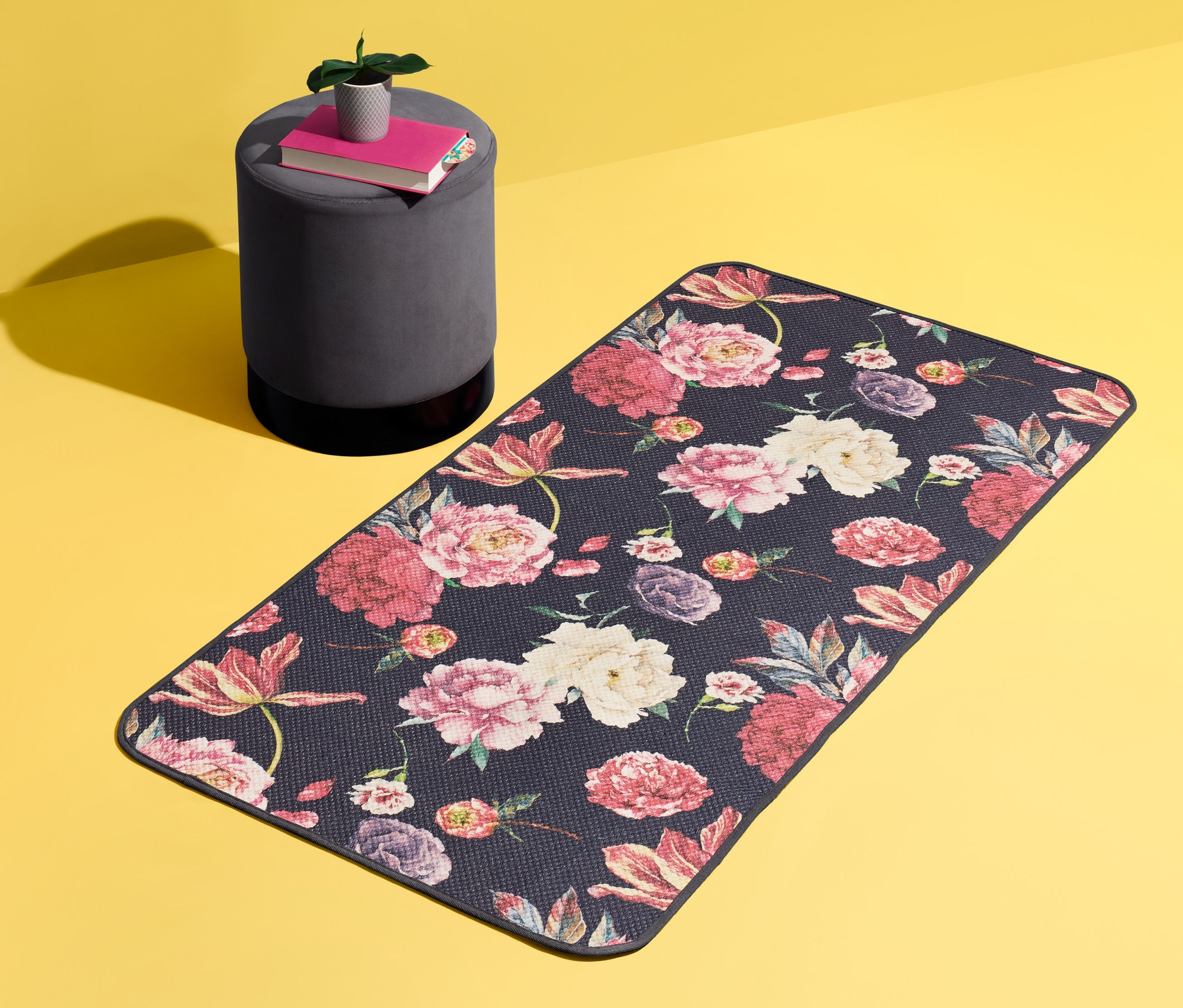 Czarny dywan w kwiaty o wymiarach ok. 140 x 70 cm leży na żółtym tle. Obok stoi szary stołek z książkami i rośliną.
