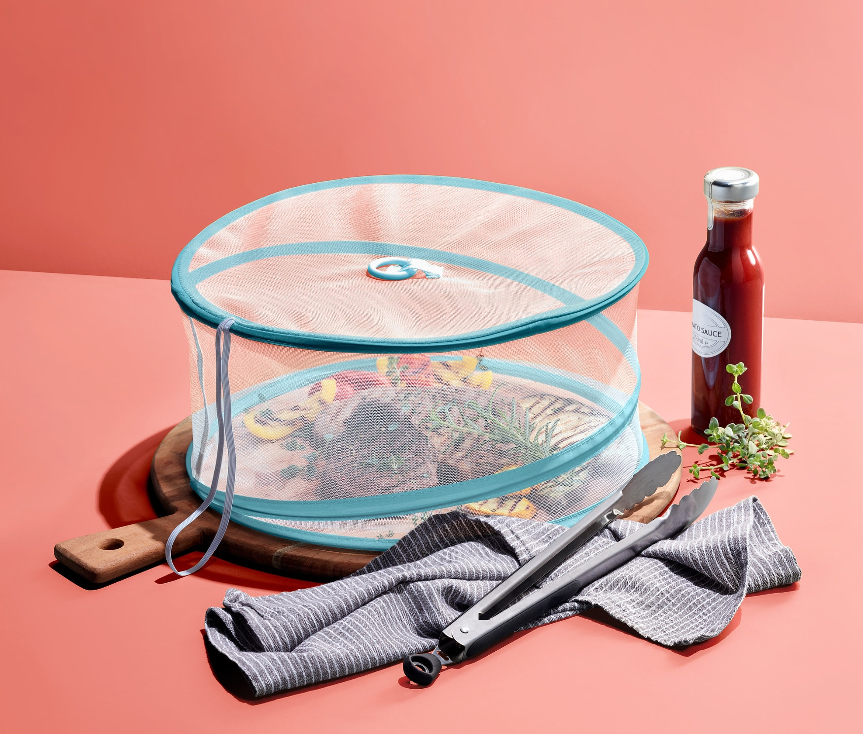 Jedzenie na drewnianej desce przykryte składaną osłoną przed owadami. Obok butelka sosu, ścierka i szczypce do grillowania.