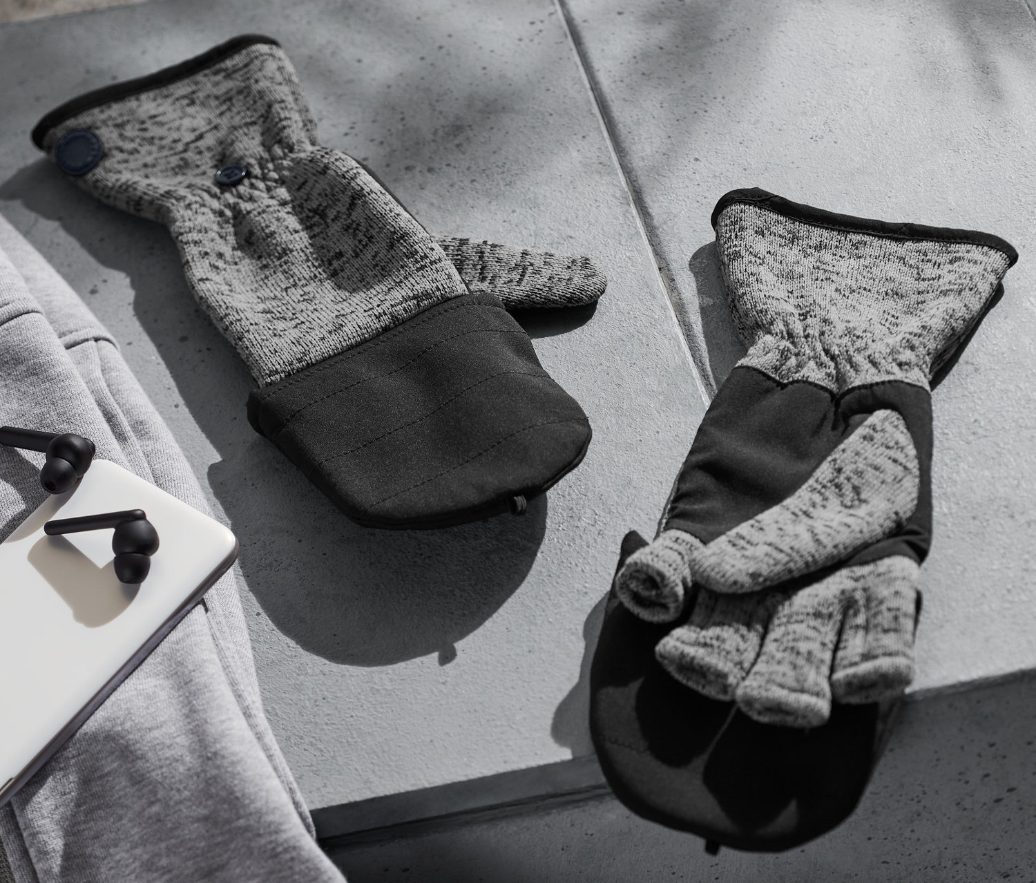 Dwie rękawice z klapką z dzianiny polarowej leżą na szarym betonowym stole obok szarej bluzy, białego telefonu i czarnych słuchawek.
