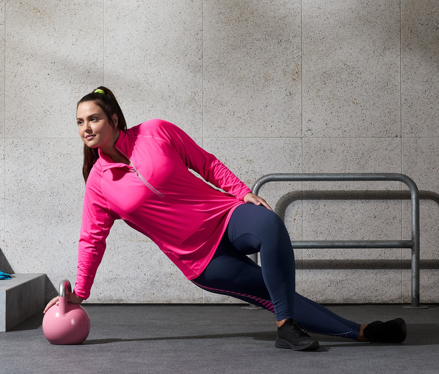 Kobieta ćwiczy z kettlebell, ma na sobie legginsy sportowe i termiczną bluzę funkcyjną, neonoworóżową.