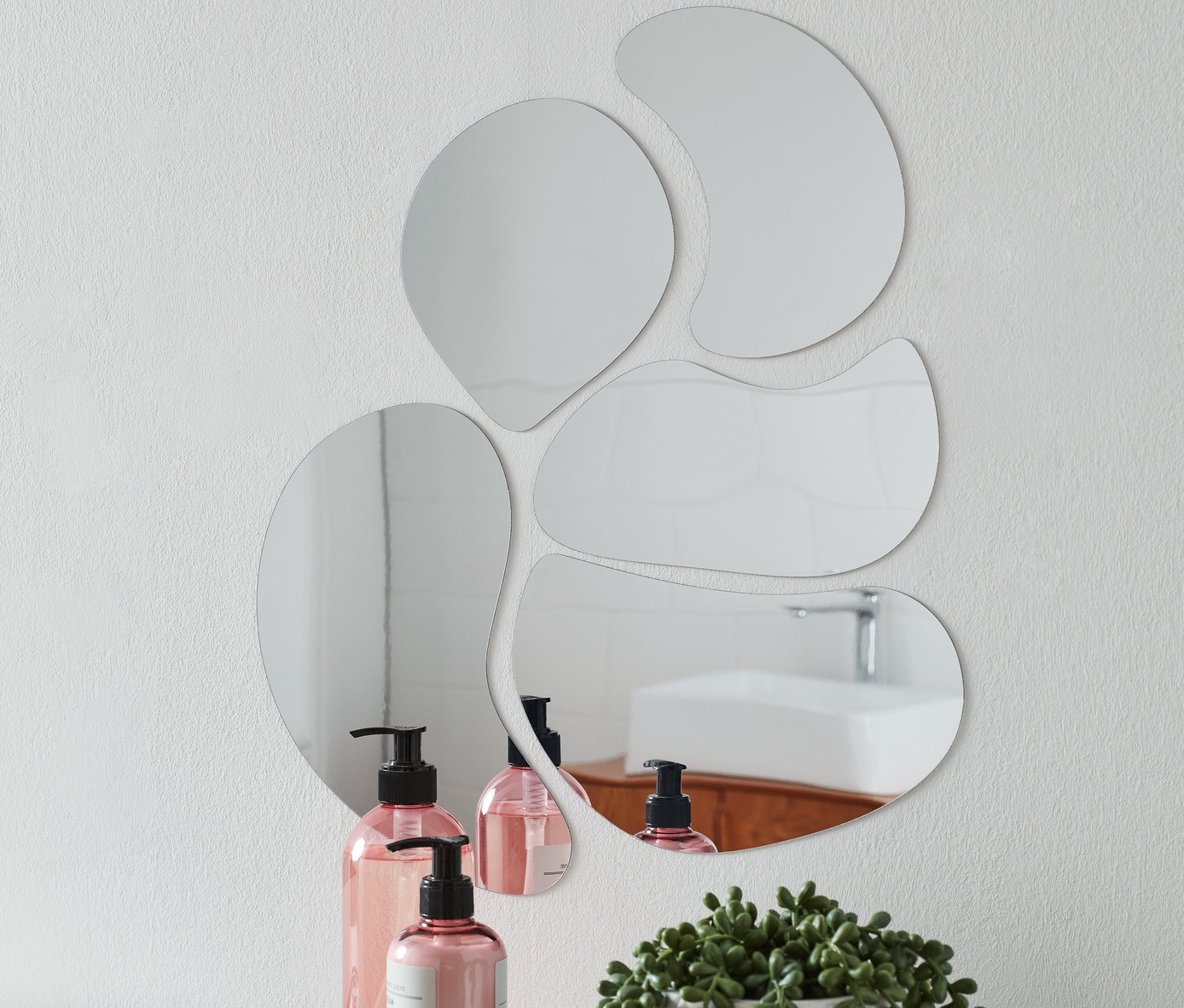 Na białej ścianie wisi pięć samoprzylepnych naklejek lustrzanych o różnych kształtach. Na białej półce pod nimi stoją dwie różowe butelki z pompką, taca na biżuterię i kwiat w białej doniczce.