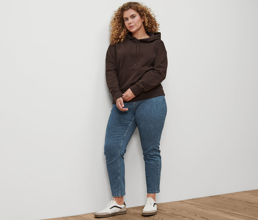 Kobieta stoi i ma na sobie jeansy Straight »Fit Juna« mid blue, skórzane sneakersy i ciemnobrązową bluzę z kapturem.