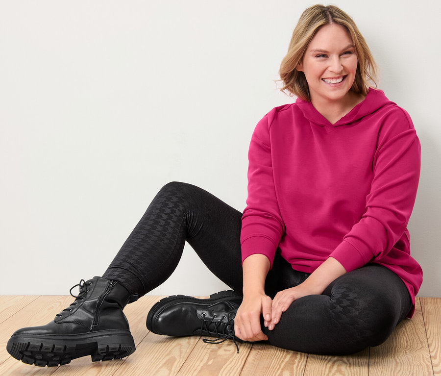 Kobieta siedzi na drewnianej podłodze, ubrana w różową bluzę z kapturem z zawartością modalu i czarne legginsy termoaktywne z tłoczeniem.