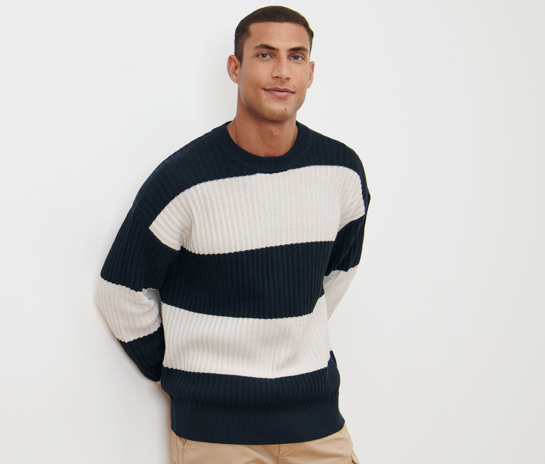 Mężczyzna pozuje na białym tle w swetrze o grubym splocie w paski granatowo-białe i spodniach khaki.