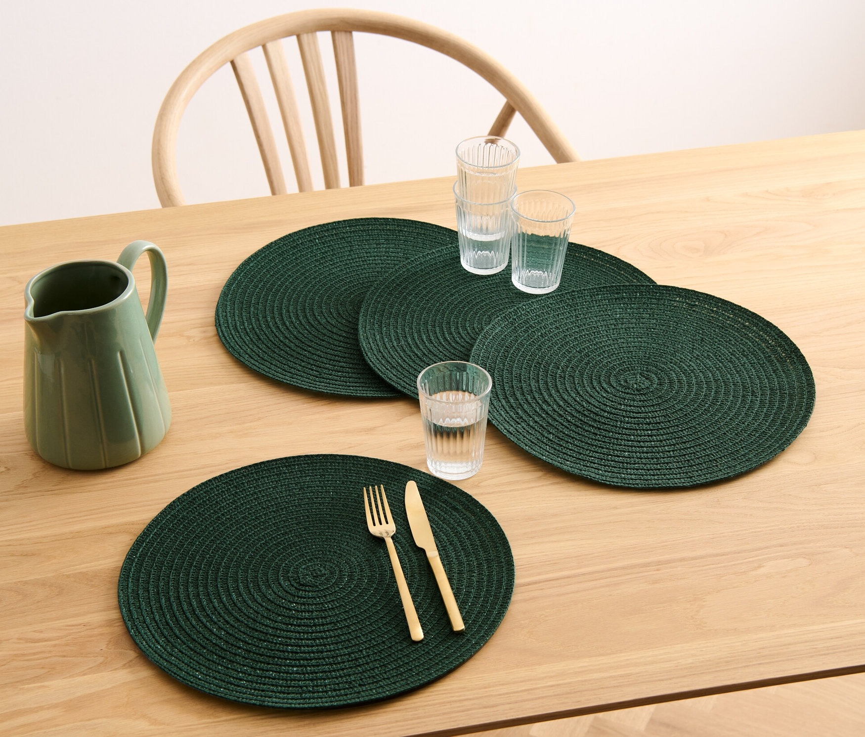 Na drewnianym stole leżą cztery zielone, okrągłe podkładki pod talerze, dzbanek i szklanki.