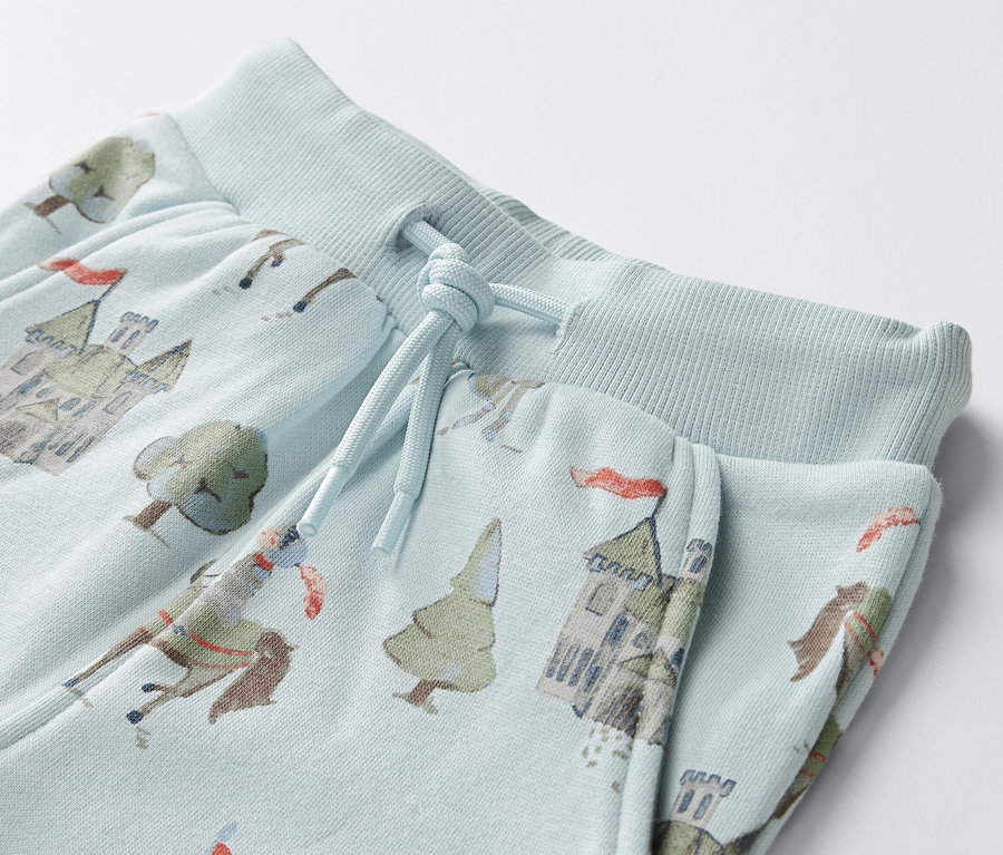 Zbliżenie na jasnoniebieskie spodnie dresowe dla niemowląt z nadrukiem rycerzy, zamków i drzew.