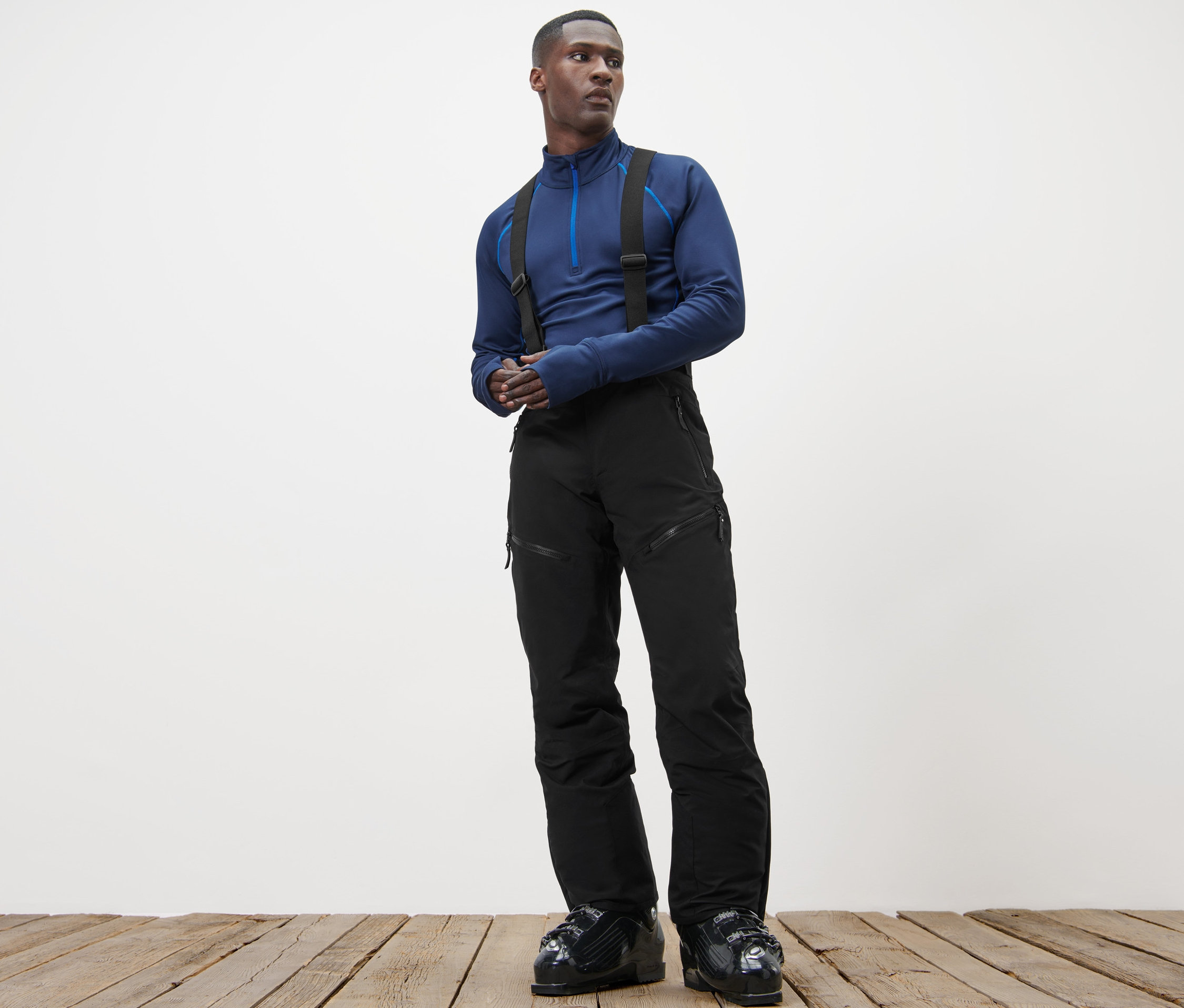 Mężczyzna stoi na drewnianej podłodze, ubrany w niebieski top i profesjonalne spodnie narciarskie.