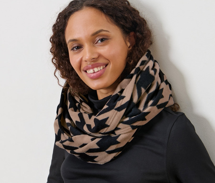 Siedząca kobieta o brązowych kręconych włosach ma na sobie wzorzysty szal komin w pepitkę.