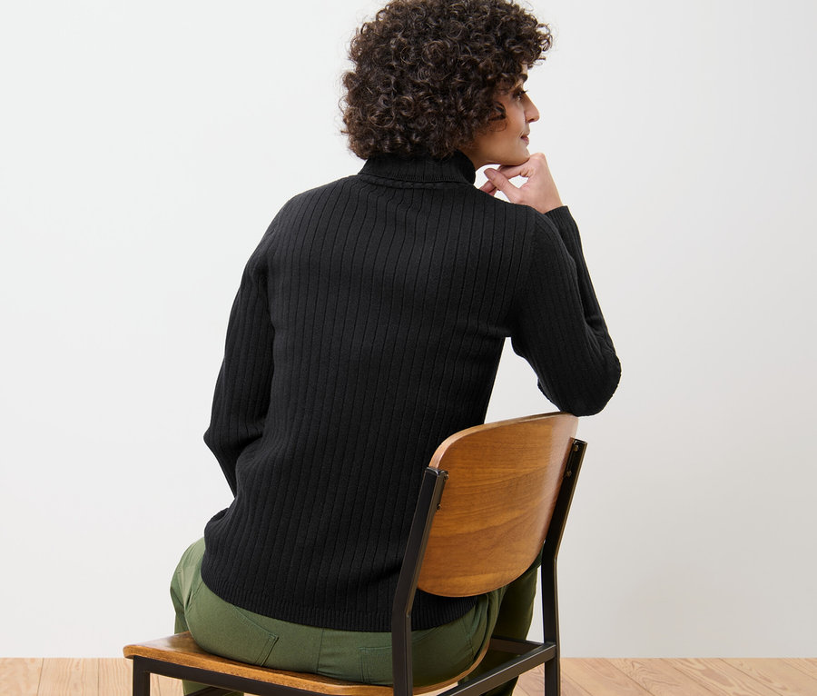 Kobieta siedzi na krześle i ma na sobie czarny golf z dzianiny o splocie ściągaczowym.