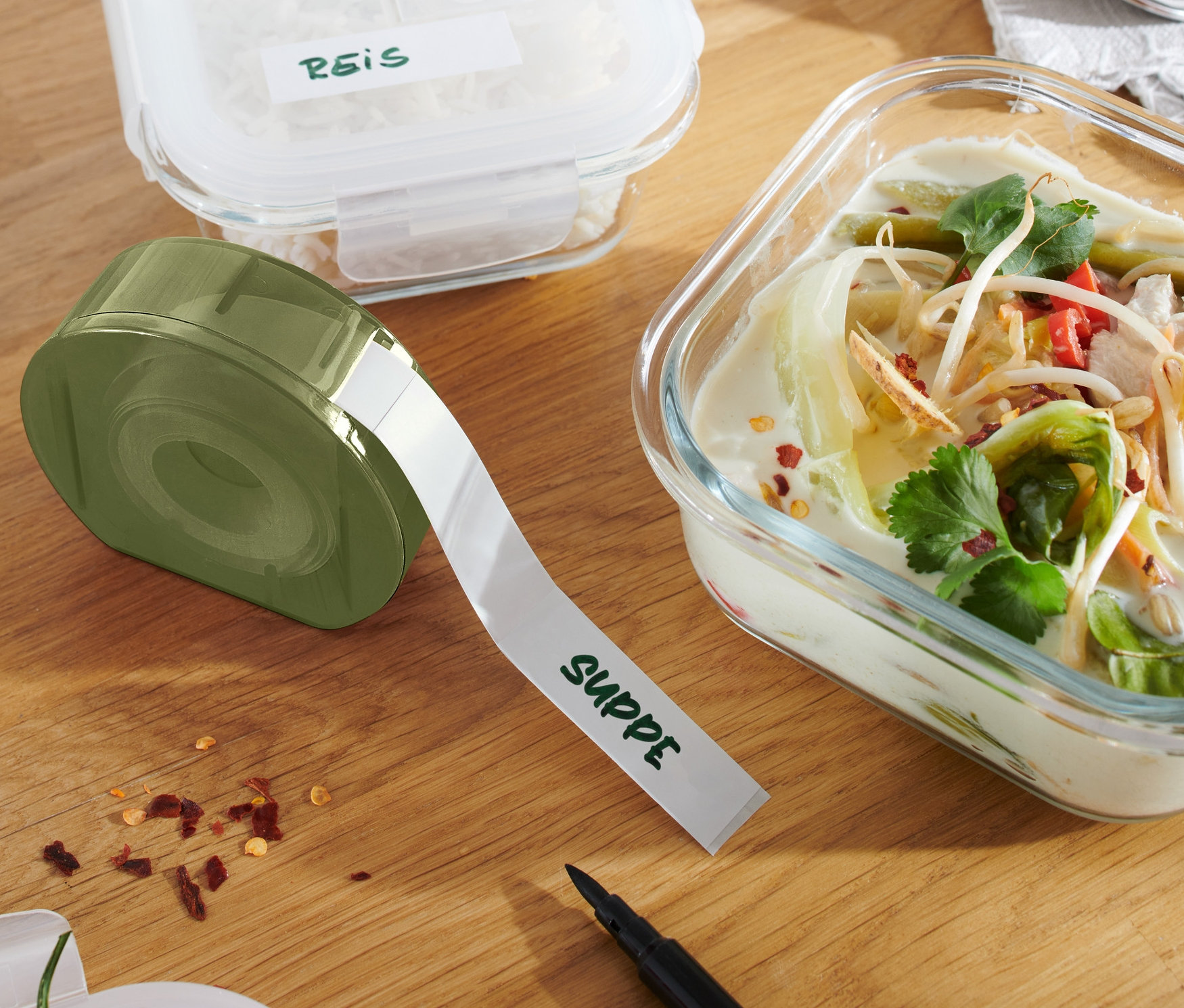 Urządzenie do etykietowania z napisem "SUPPE" leży obok szklanego pojemnika z zupą i innego pojemnika z ryżem.