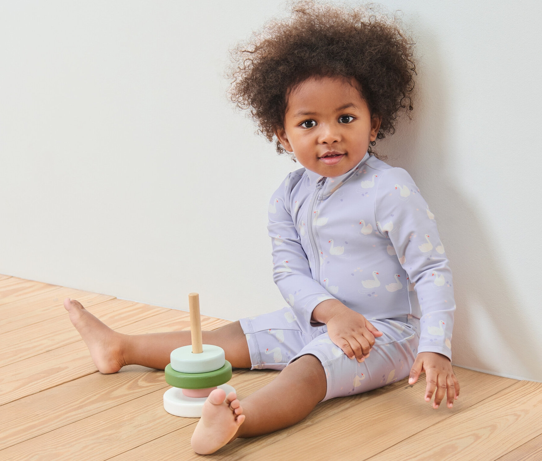 Dziecko siedzi na drewnianej podłodze obok drewnianej zabawki. Ma na sobie kombinezon niemowlęcy z filtrem UV 80, łabędź.