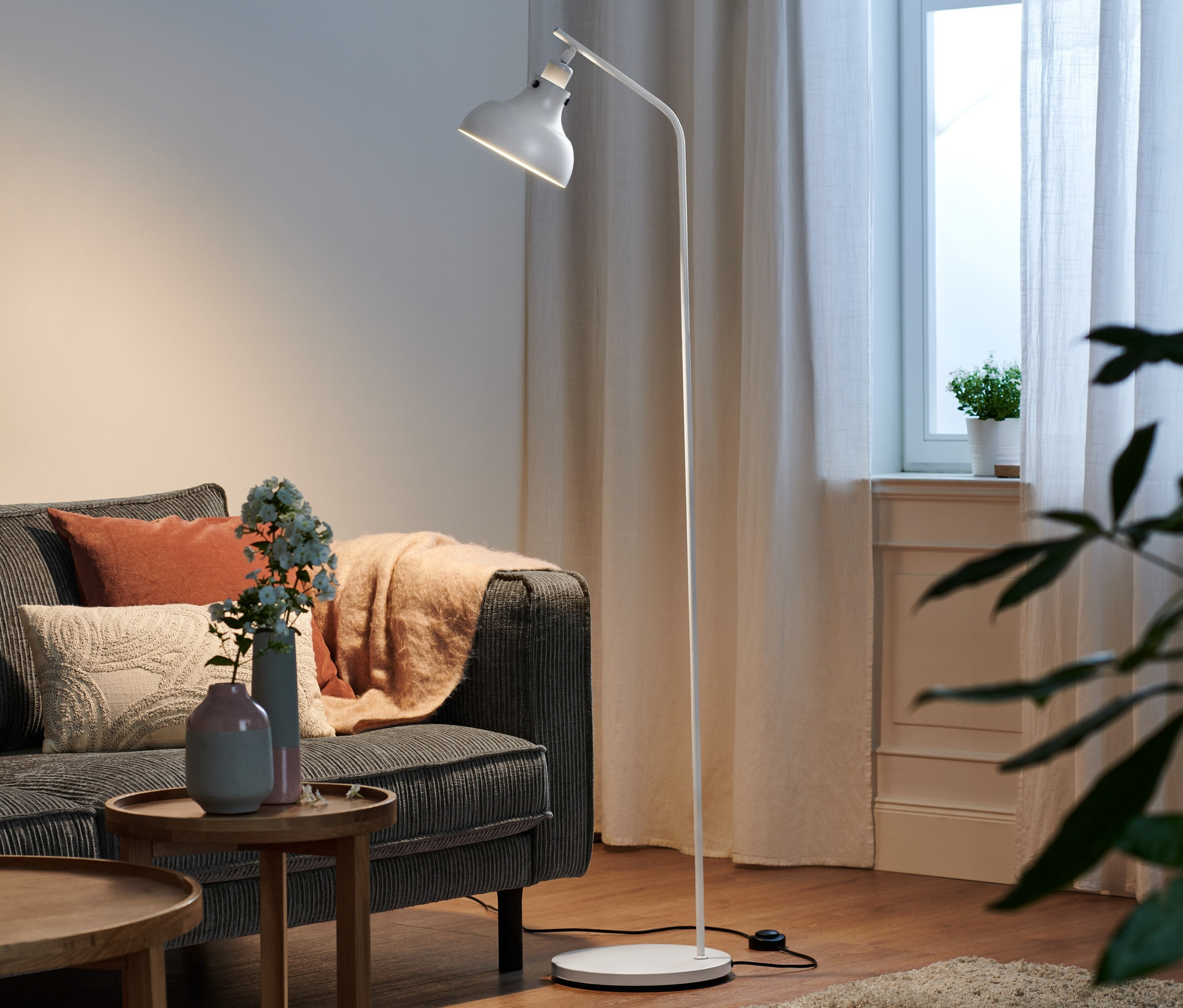 W salonie stoi metalowa lampa stojąca obok szarej sofy.