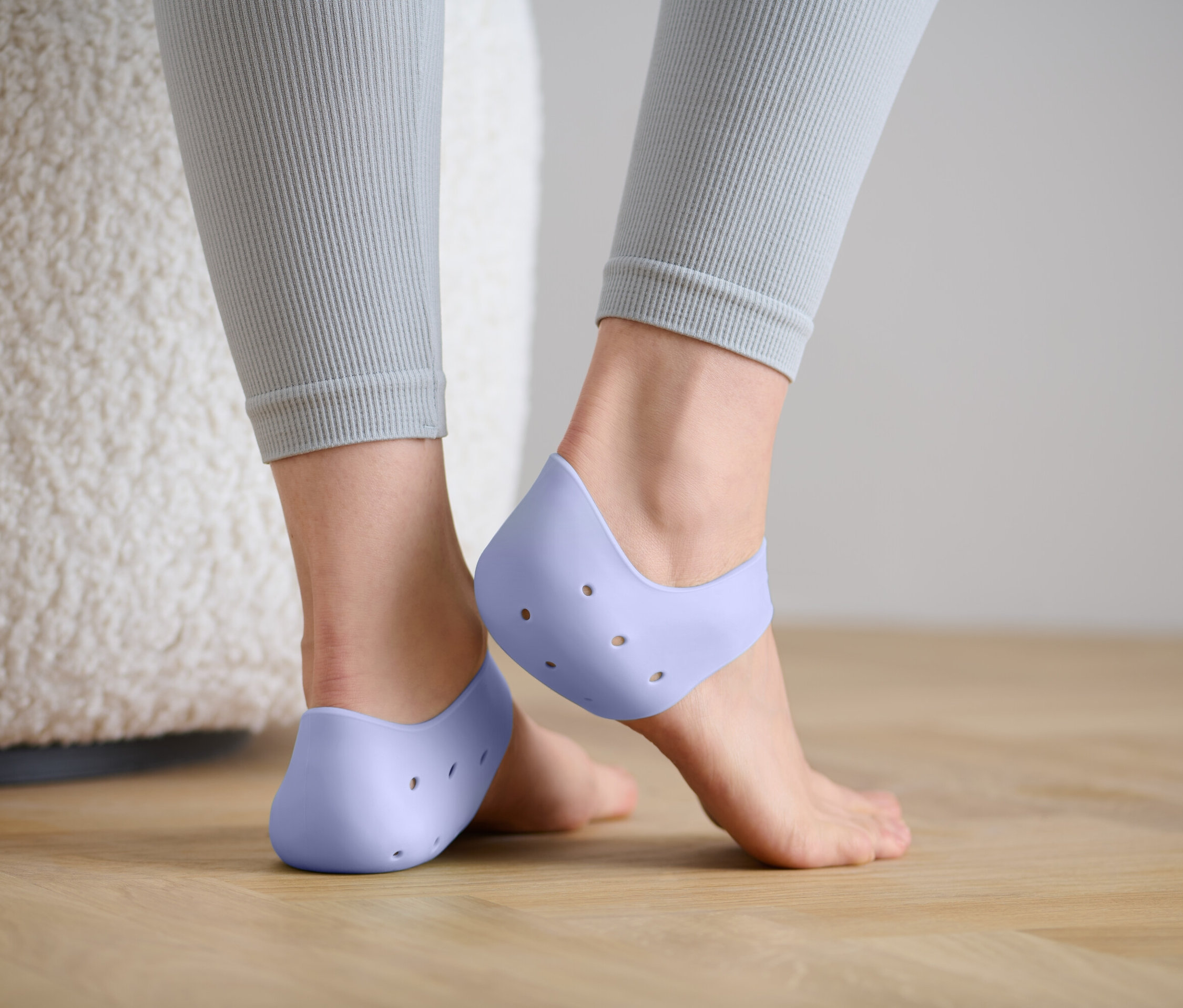 Widok z tyłu kobiety w szarych legginsach, która ma na stopach jasno fioletowe silikonowe ochraniacze na pięty z otworami.