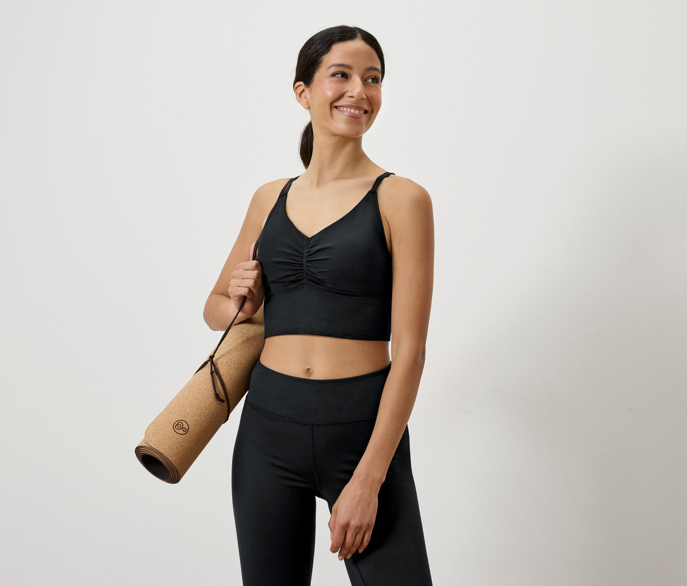 Uśmiechnięta kobieta w czarnym stroju sportowym trzyma pod pachą zwiniętą matę do jogi.