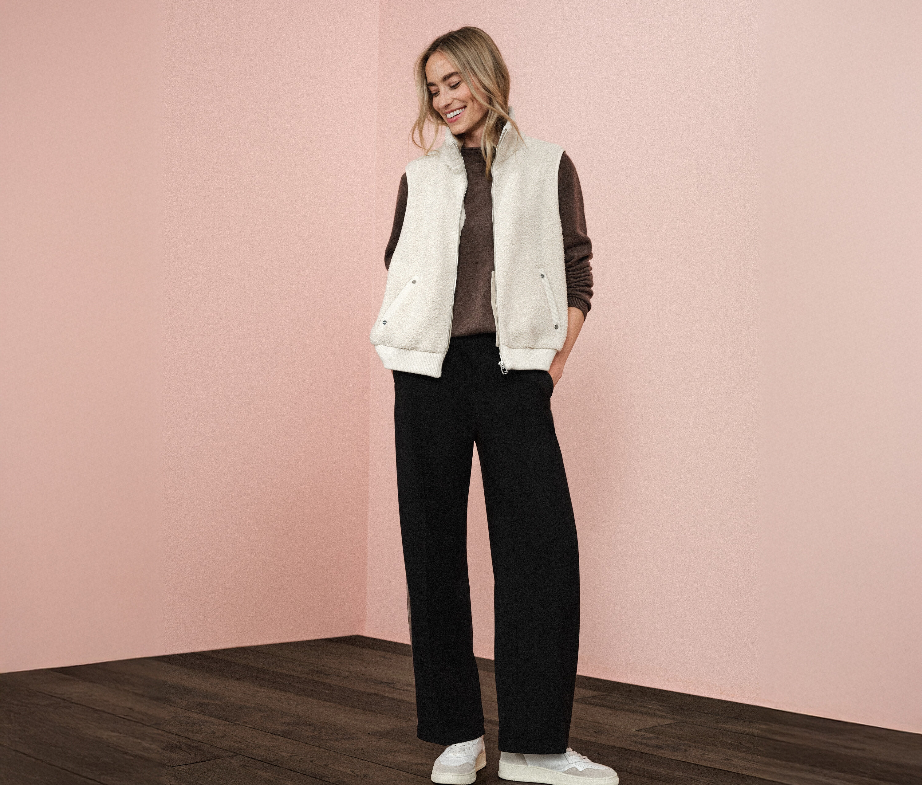 Kobieta nosi pluszową kamizelkę Street One na brązowym swetrze, w połączeniu z czarnymi spodniami i białymi butami sportowymi.