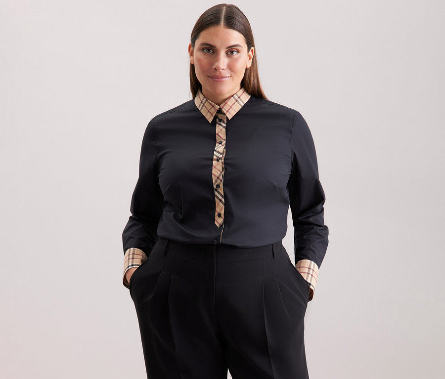 Kobieta o brązowych włosach ma na sobie czarną popelinową bluzkę Seidensticker z detalami w kratę i czarne spodnie.