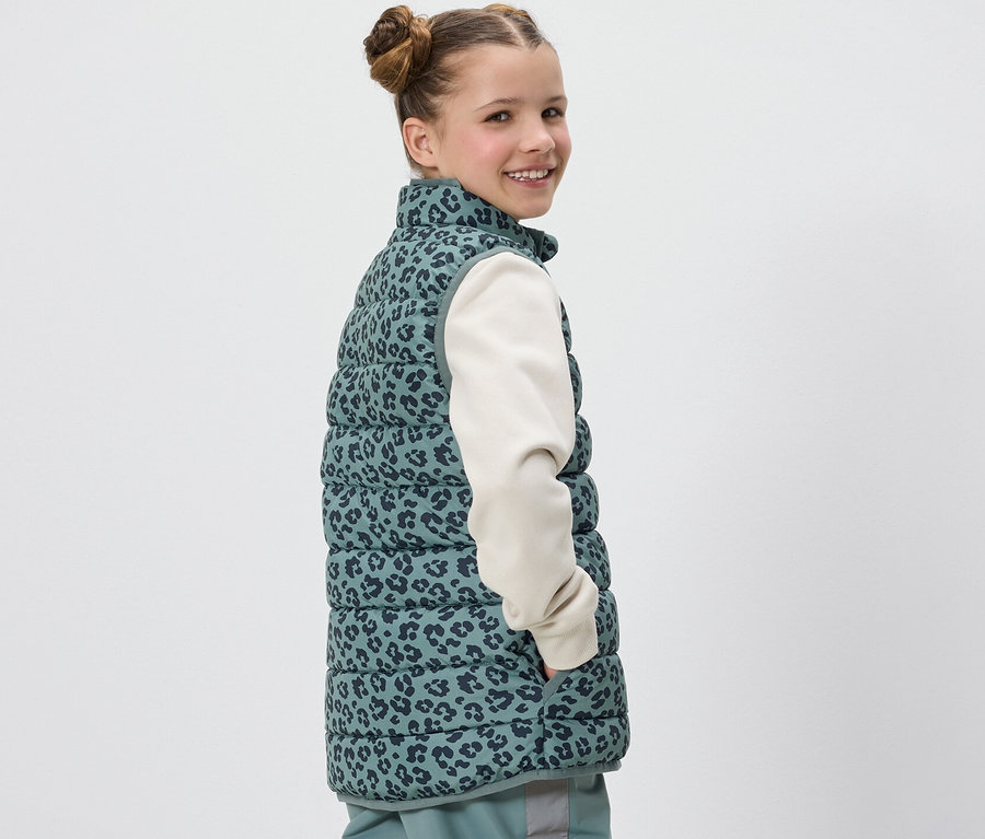 Dziewczyna uśmiecha się przez ramię, ubrana w dziecięcą kamizelkę pikowaną w panterkę.