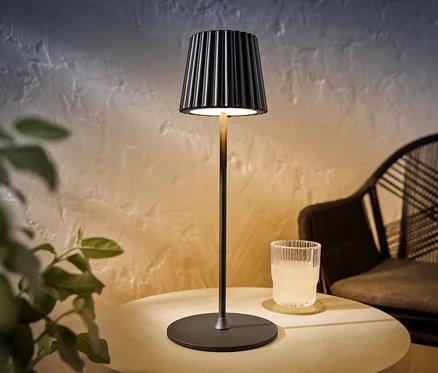 Czarna akumulatorowa lampa stołowa LED stoi na stole obok szklanki z napojem.
