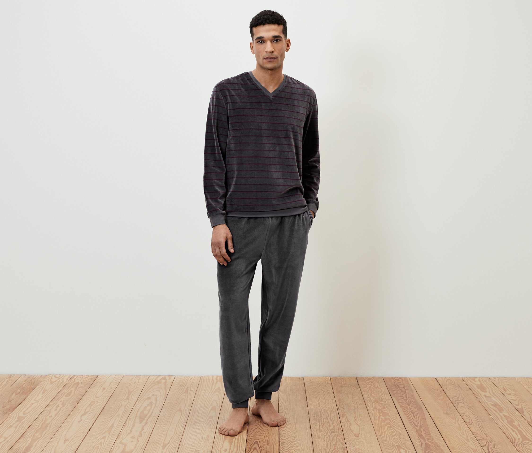 Mężczyzna stoi boso, ubrany w szarą, prążkowaną piżamę welurową.