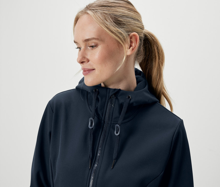 Portret kobiety z blond włosami związanymi w kucyk, ubranej w granatową bluzę z kapturem zapinaną na zamek.