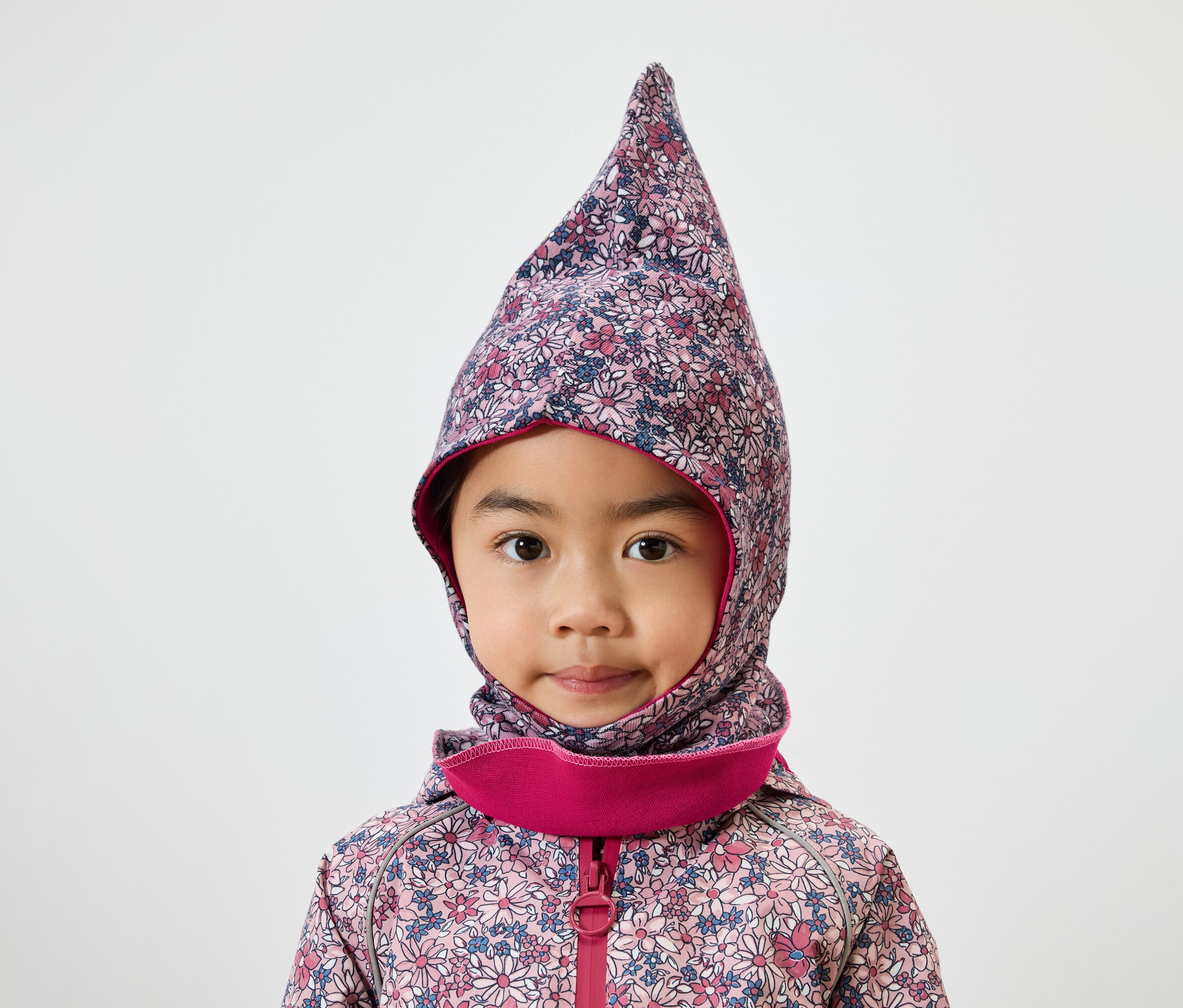 Portret małej dziewczynki o brązowych oczach w kwiatowej kominiarce z różowym obszyciem i kurtce.