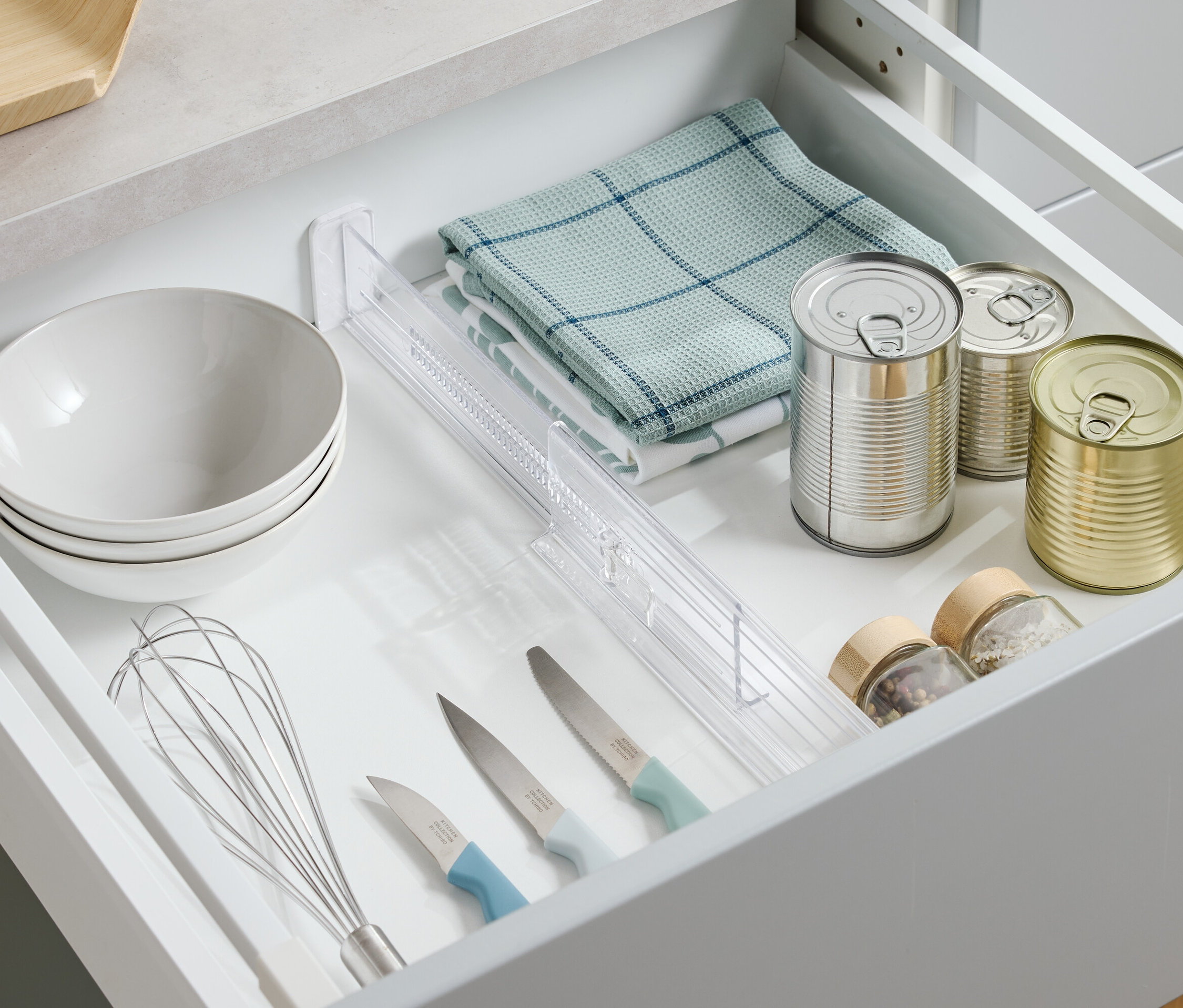 Widok z góry na otwartą szufladę z miskami, ściereczką, puszkami, przyprawami, nożami i trzepaczką.