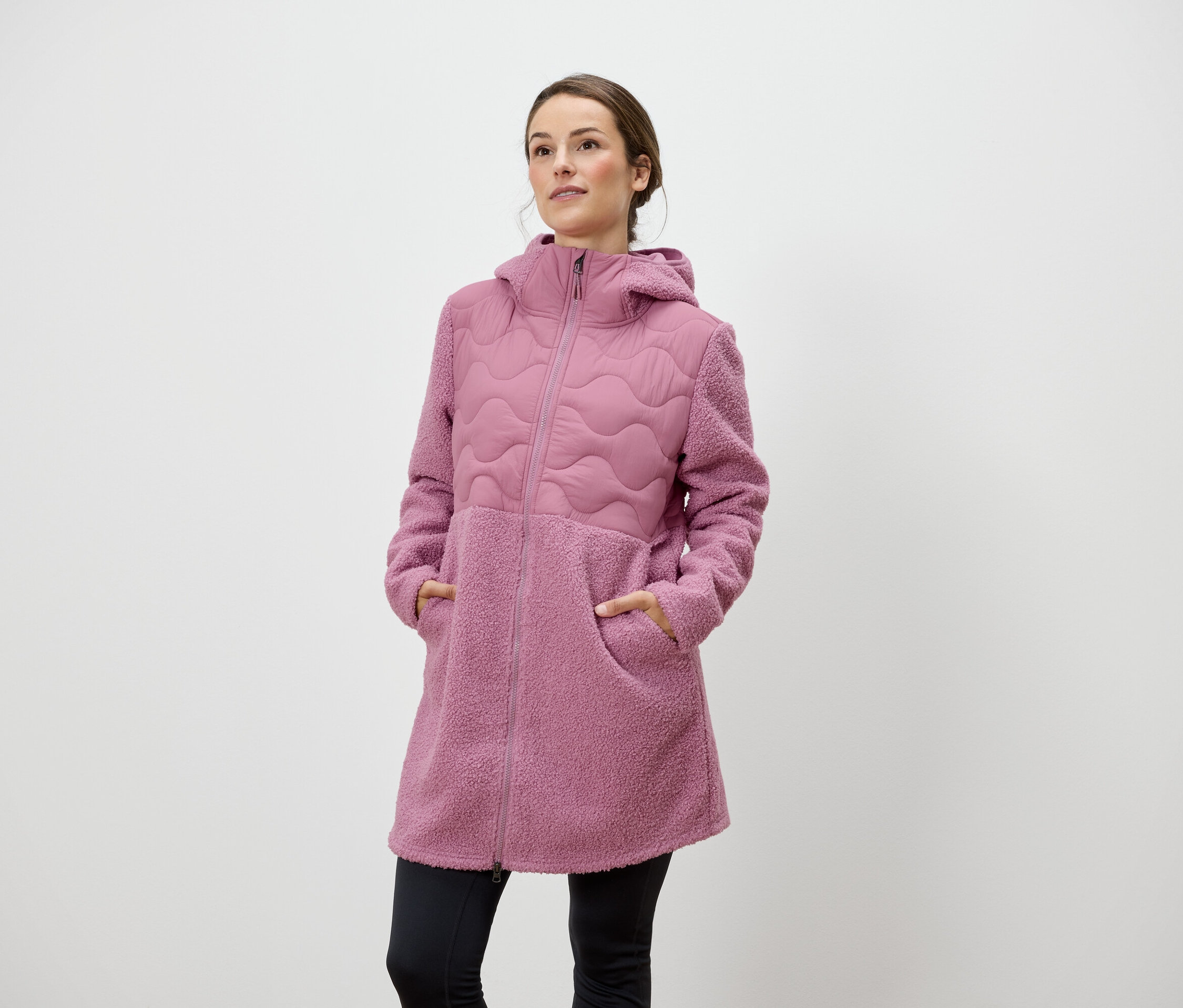 Kobieta stoi z rękami w kieszeniach różowego płaszcza zapinanego na zamek z kapturem.